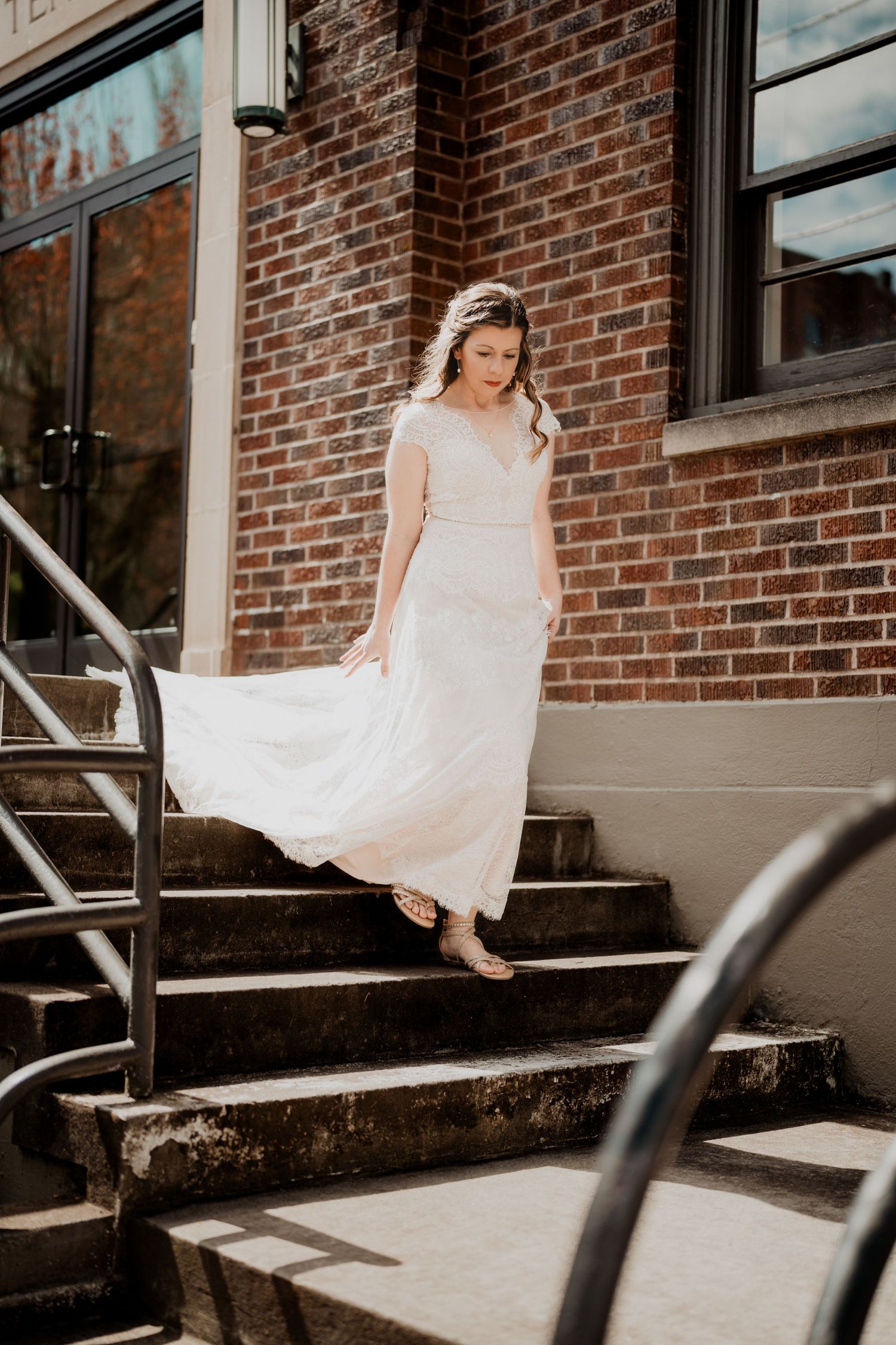 Bride poses gracefully on concrete steps against classic brick building facade.
