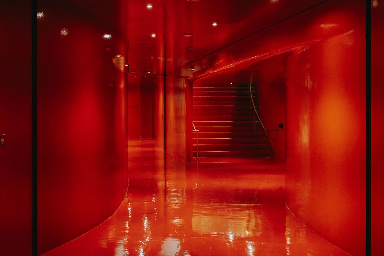 A sleek red corridor with reflective floors leads to a staircase, bathed in dramatic crimson lighting.