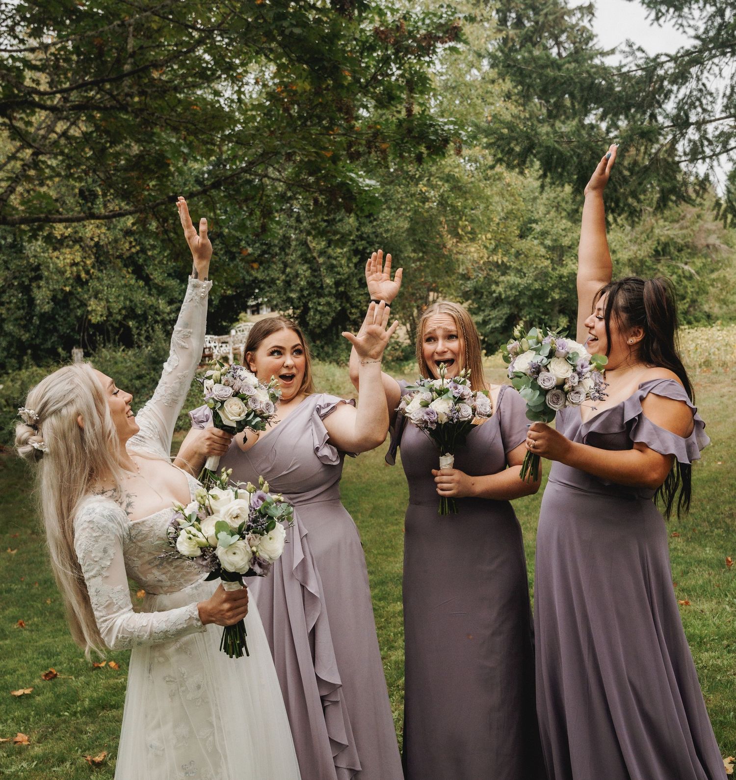 Wedding party wearing dusty purple dresses celebrate together in a joyful group photo outdoors in a garden setting.