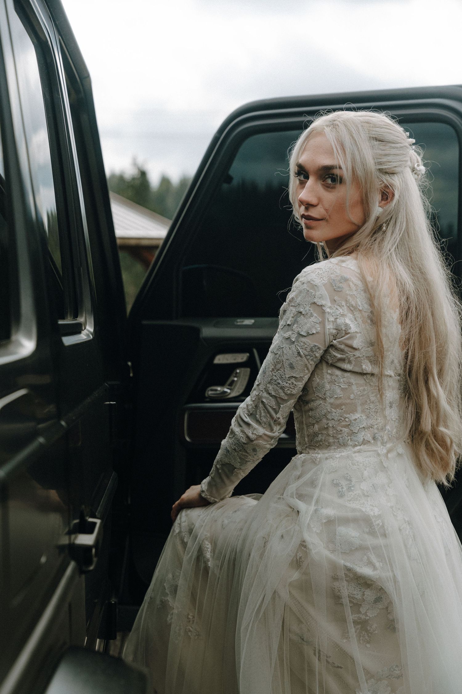 A bride in a lace and tulle wedding dress poses beside a black car door.