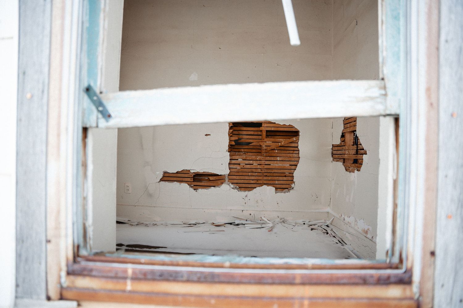 View through white window frame showing damaged drywall with exposed wood lath underneath.