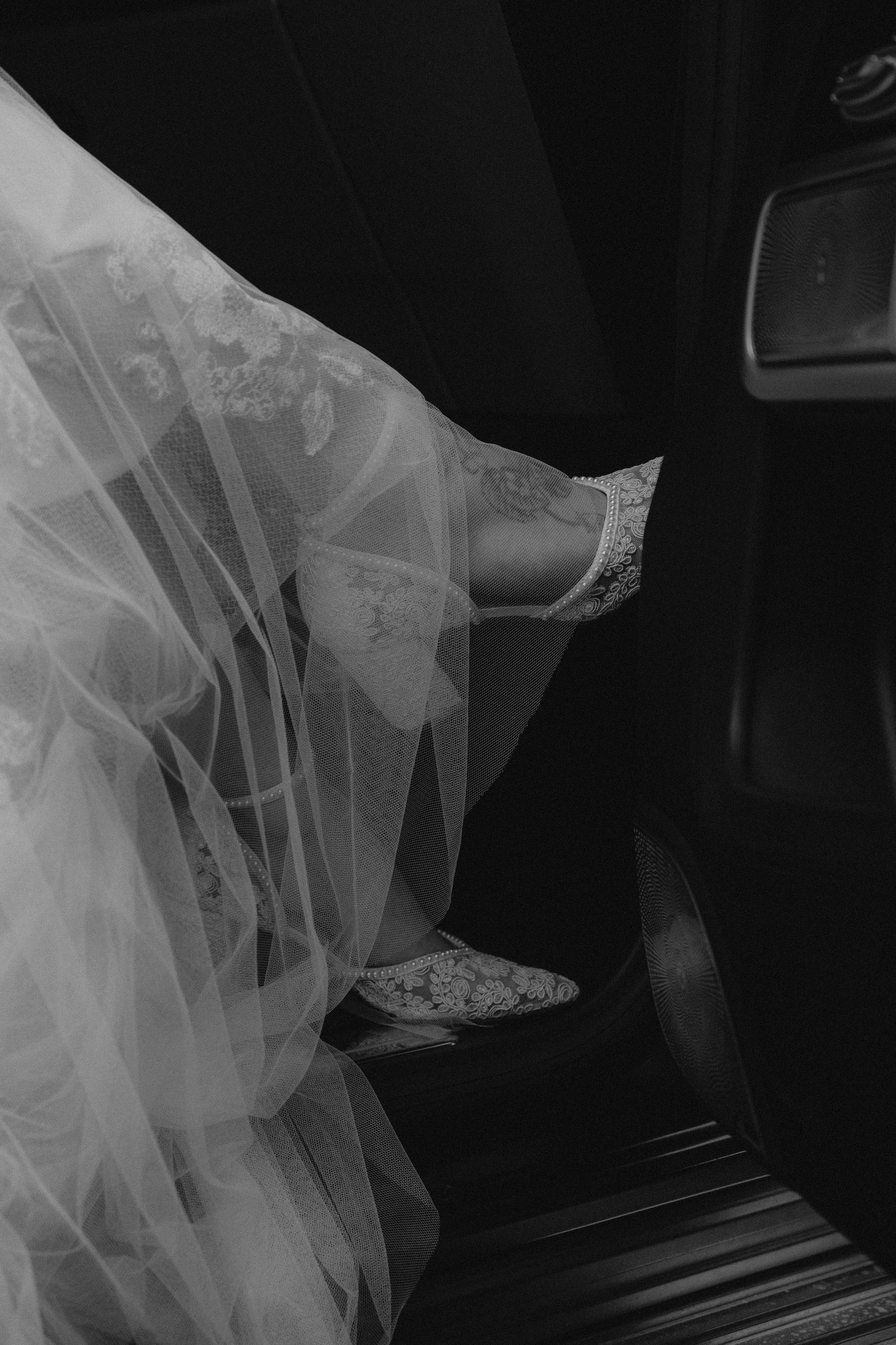 A close-up black and white photo of ornate bridal shoes with lace and tulle details in a car.