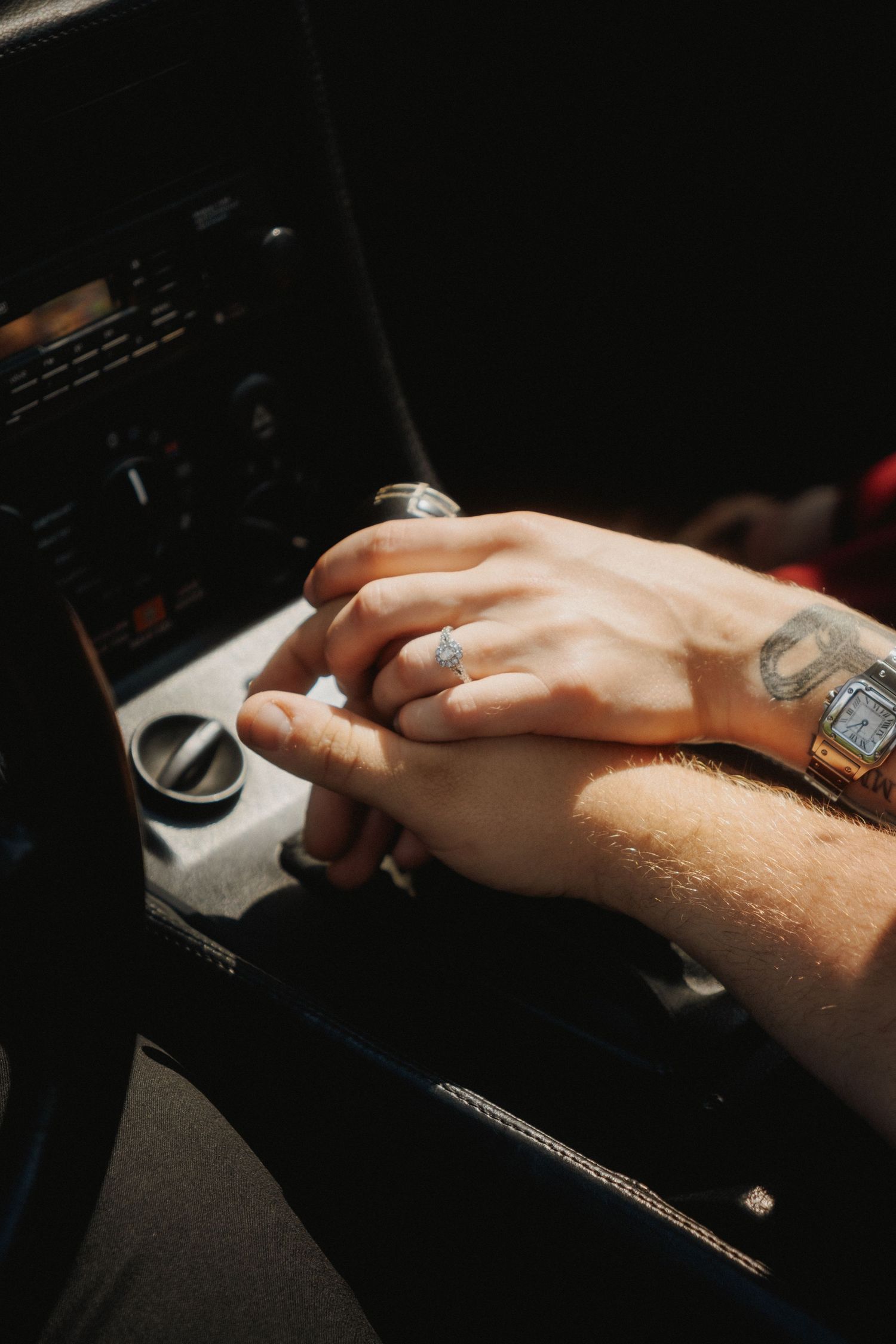 Close-up detail of wedding rings and hands on car console showing tattoo and watch accessories.