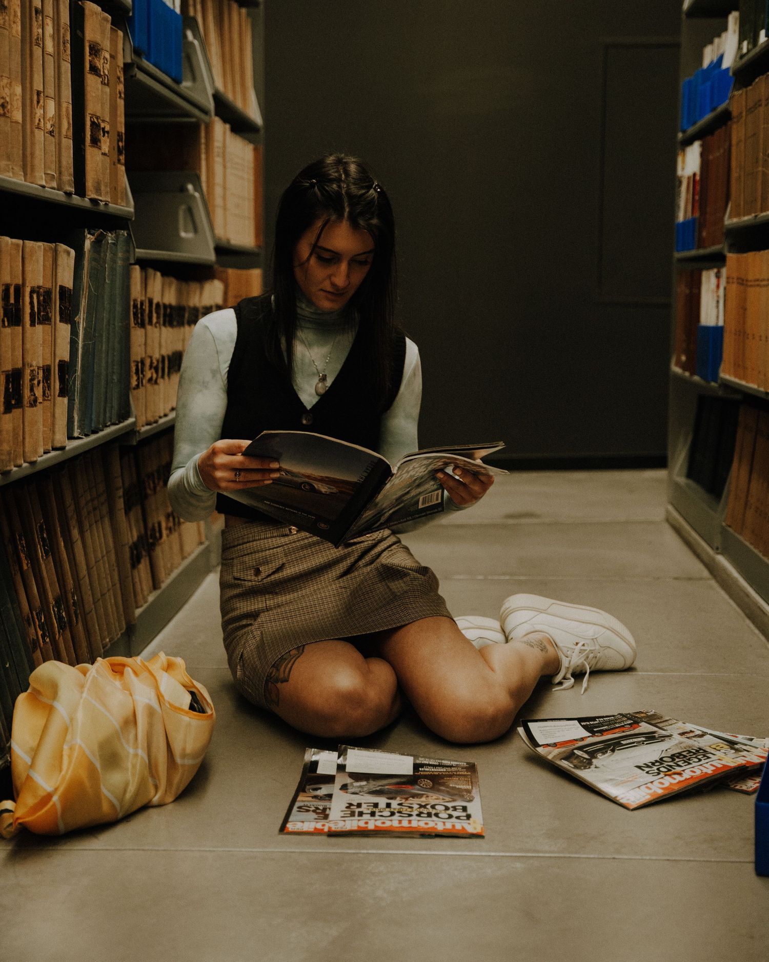 Someone sits on a library floor between bookshelves reading magazines with a yellow backpack nearby.