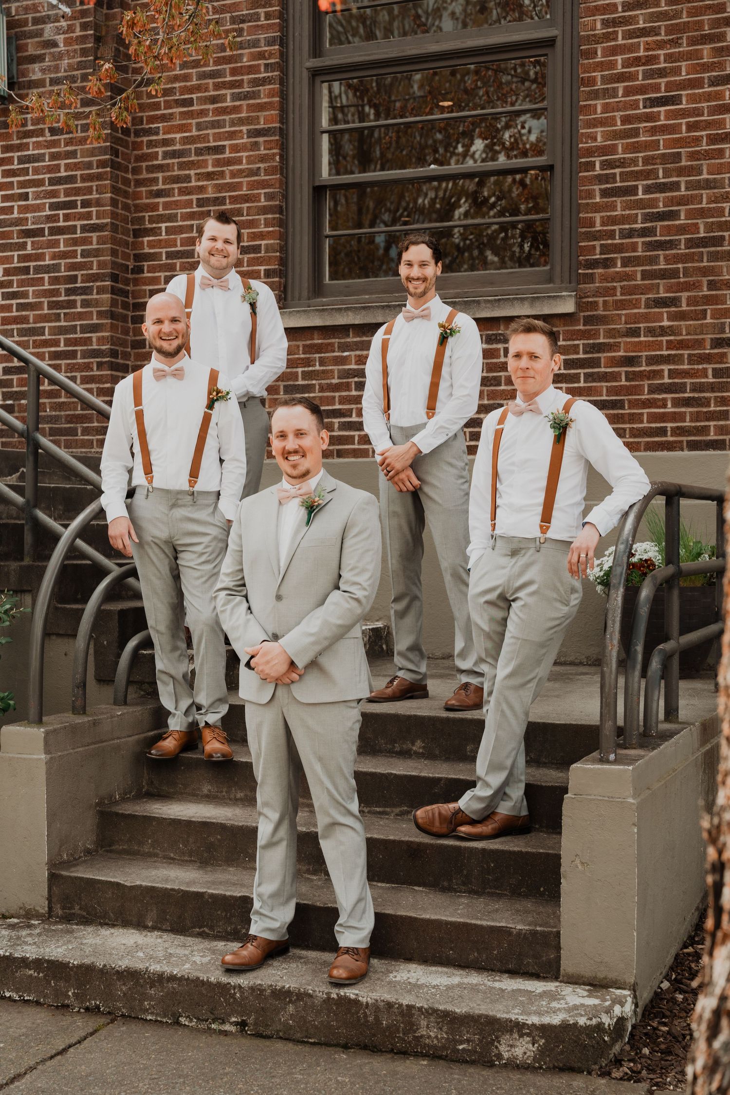 Groomsmen in light grey suits with brown suspenders pose on brick building steps.
