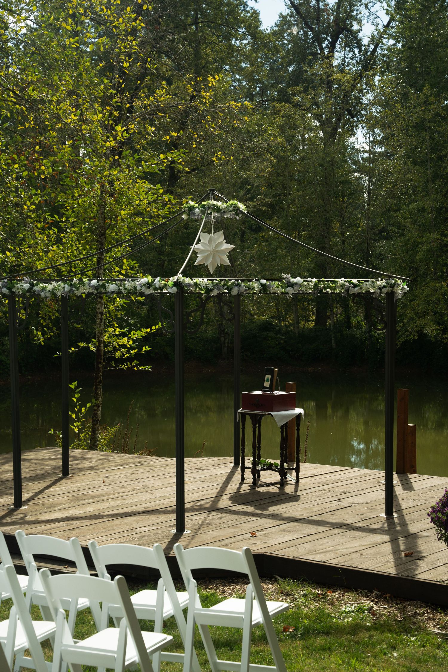 Outdoor wedding ceremony setup with white chairs and a canopy overlooking a serene pond in a wooded setting.