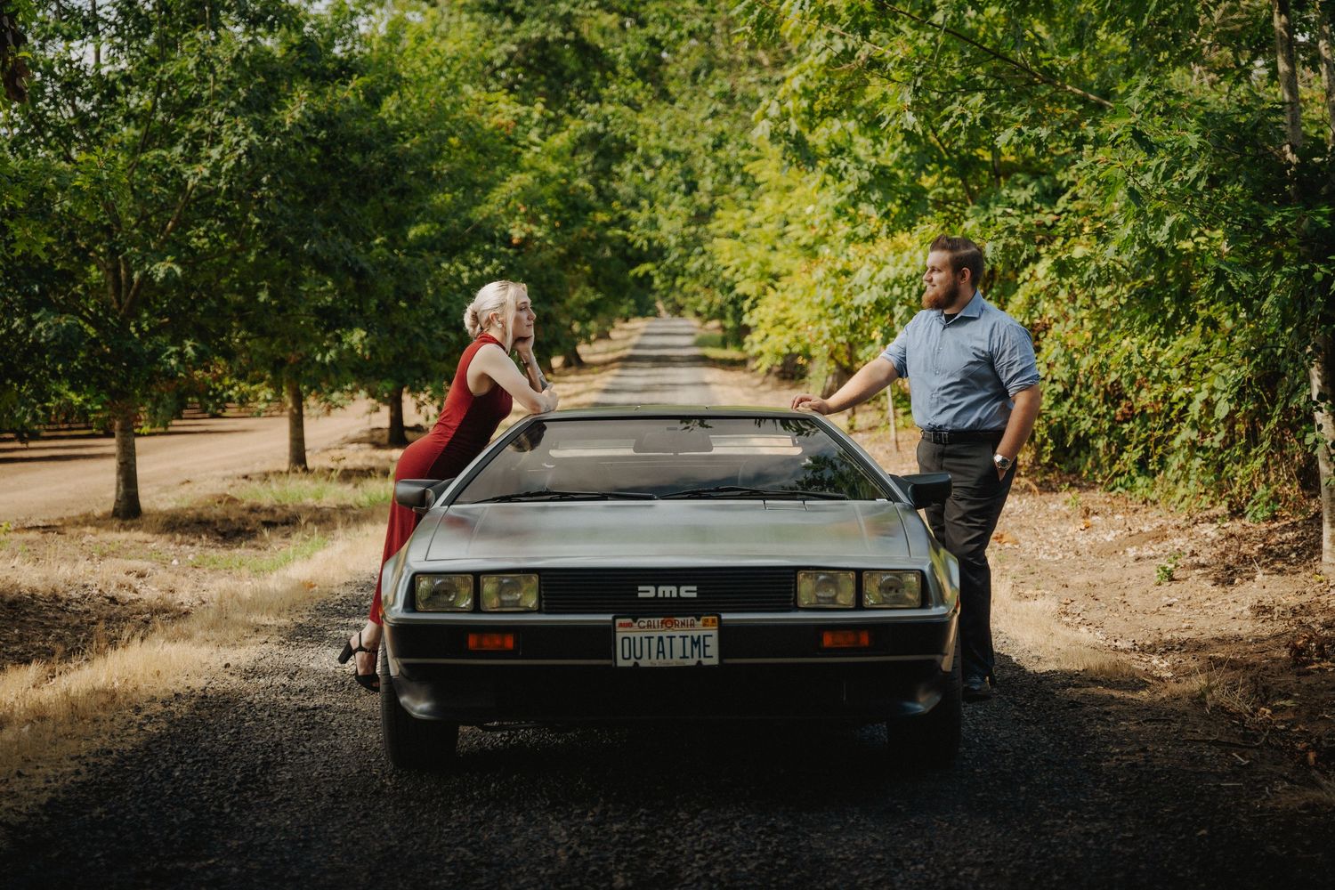 A classic DeLorean DMC-12 sports car parked on a tree-lined path with its iconic gull-wing doors raised.