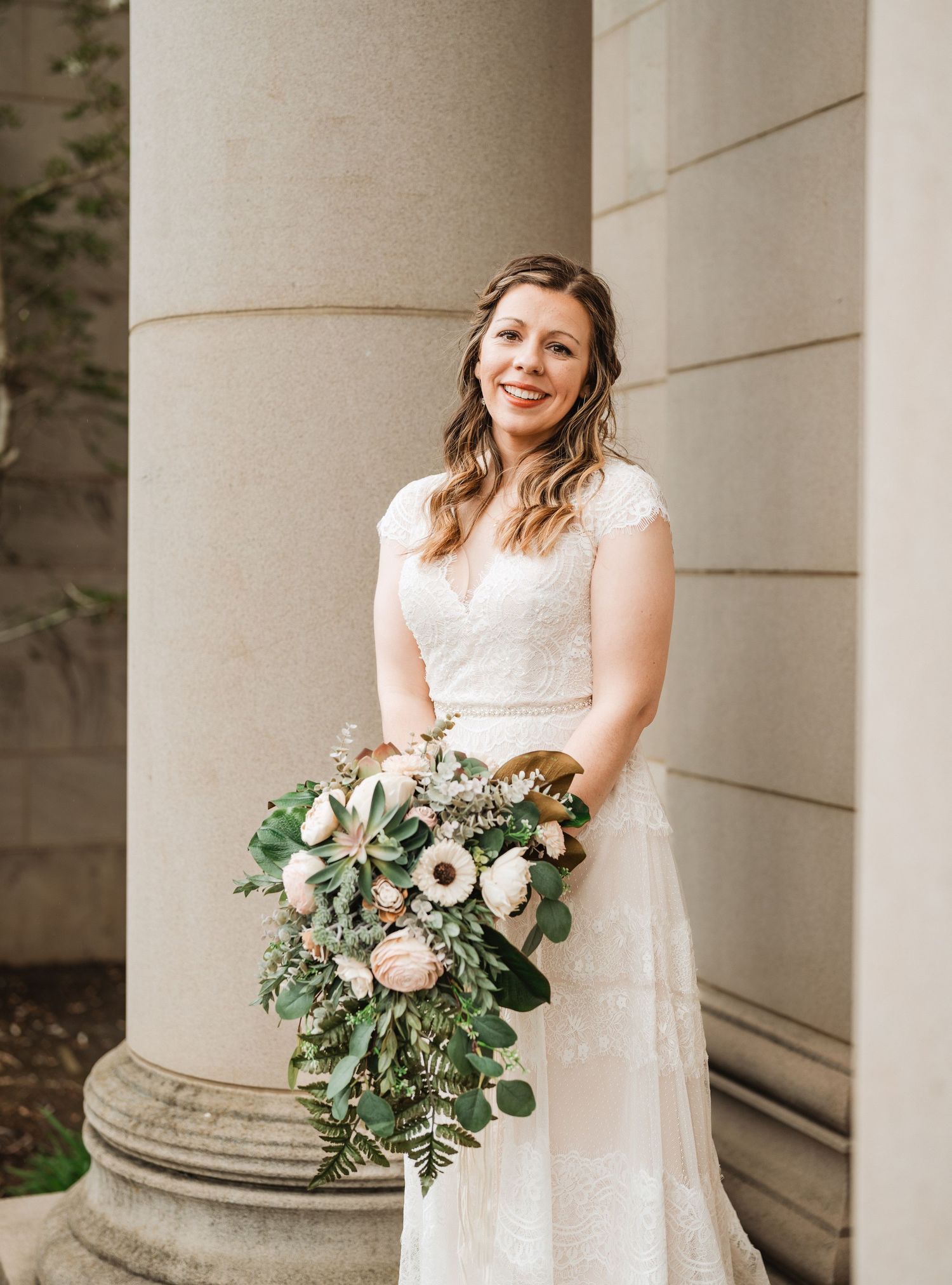 Beautiful bride in lace cap sleeve wedding dress holds lush green and white cascading bouquet against stone column.
