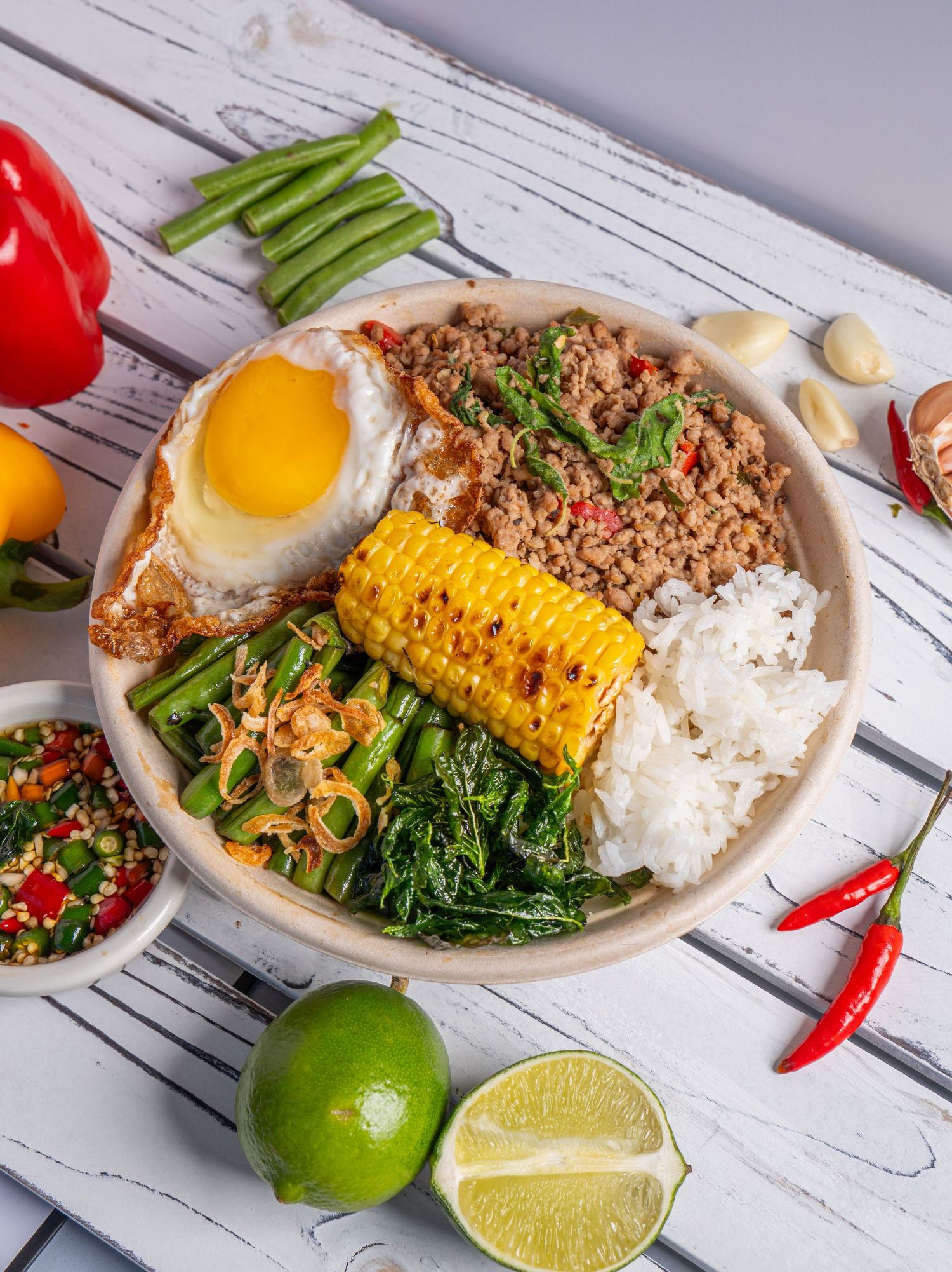 Vibrant rice bowl with minced meat, fried egg, grilled corn, sauteed greens, served with lime and chili peppers on white wood.