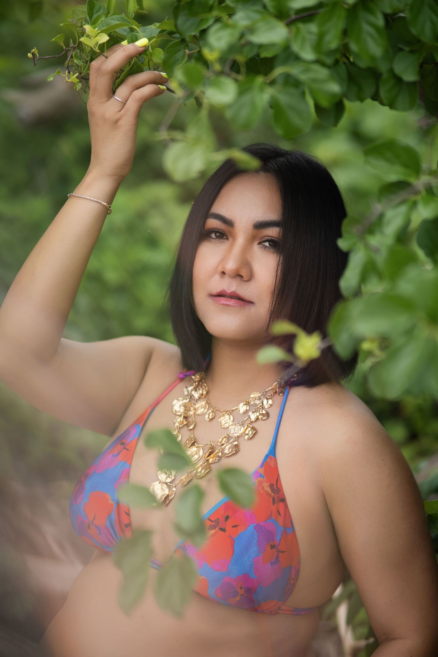 Woman in floral print top and gold necklace poses against lush green foliage in outdoor portrait setting.