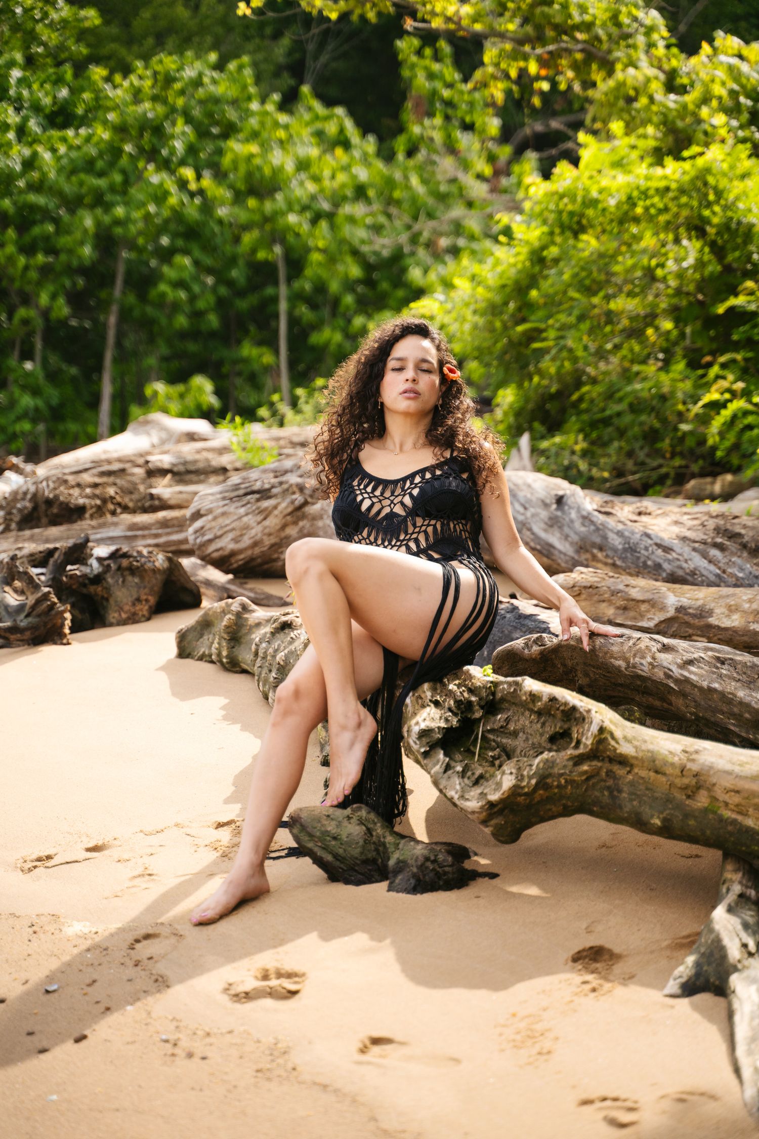 A person in black attire poses on driftwood at a sandy beach with lush greenery in the background.