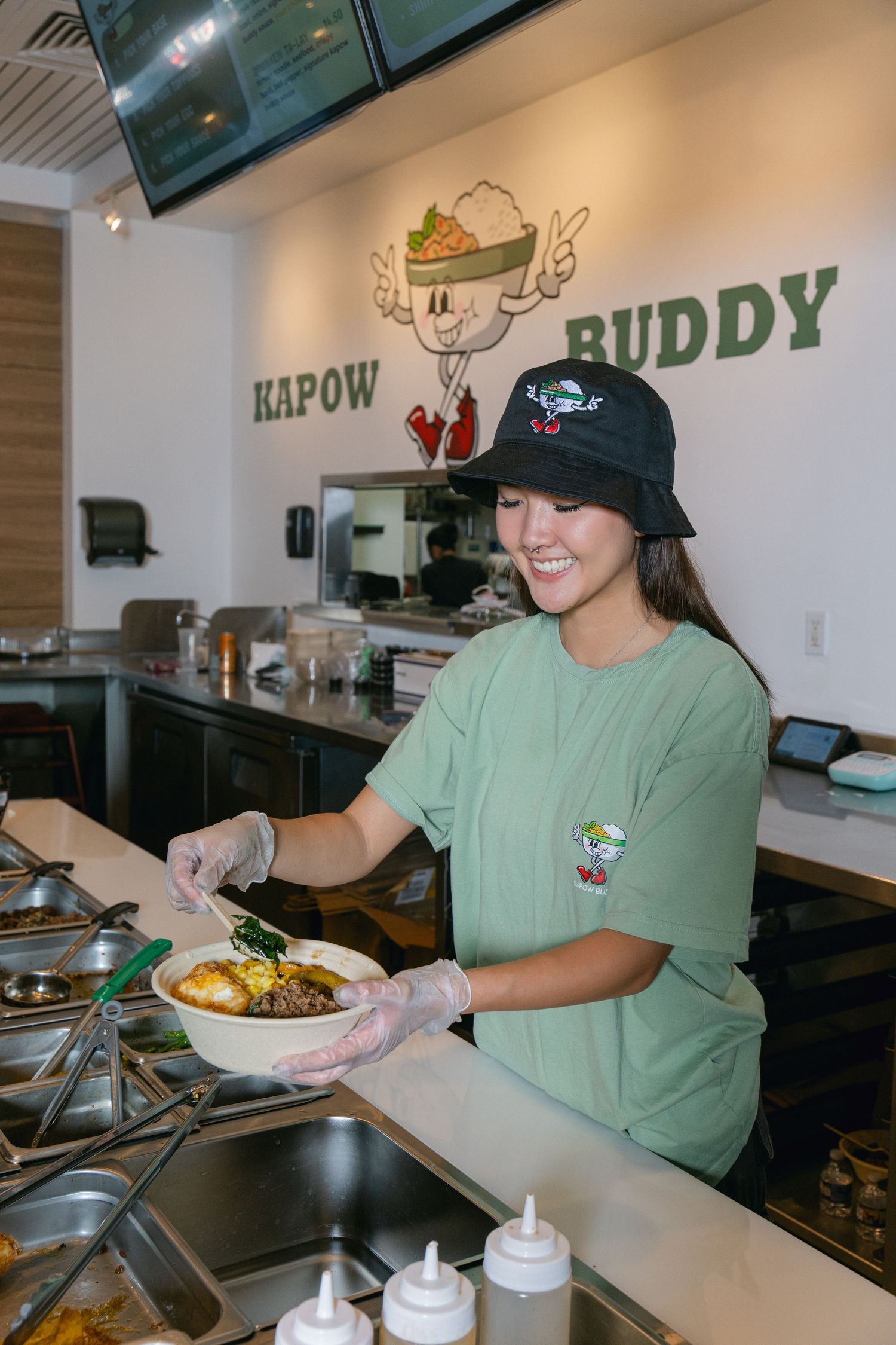 Restaurant worker in a green t-shirt and black cap plates food at Kapow Buddy counter with logo visible on wall.