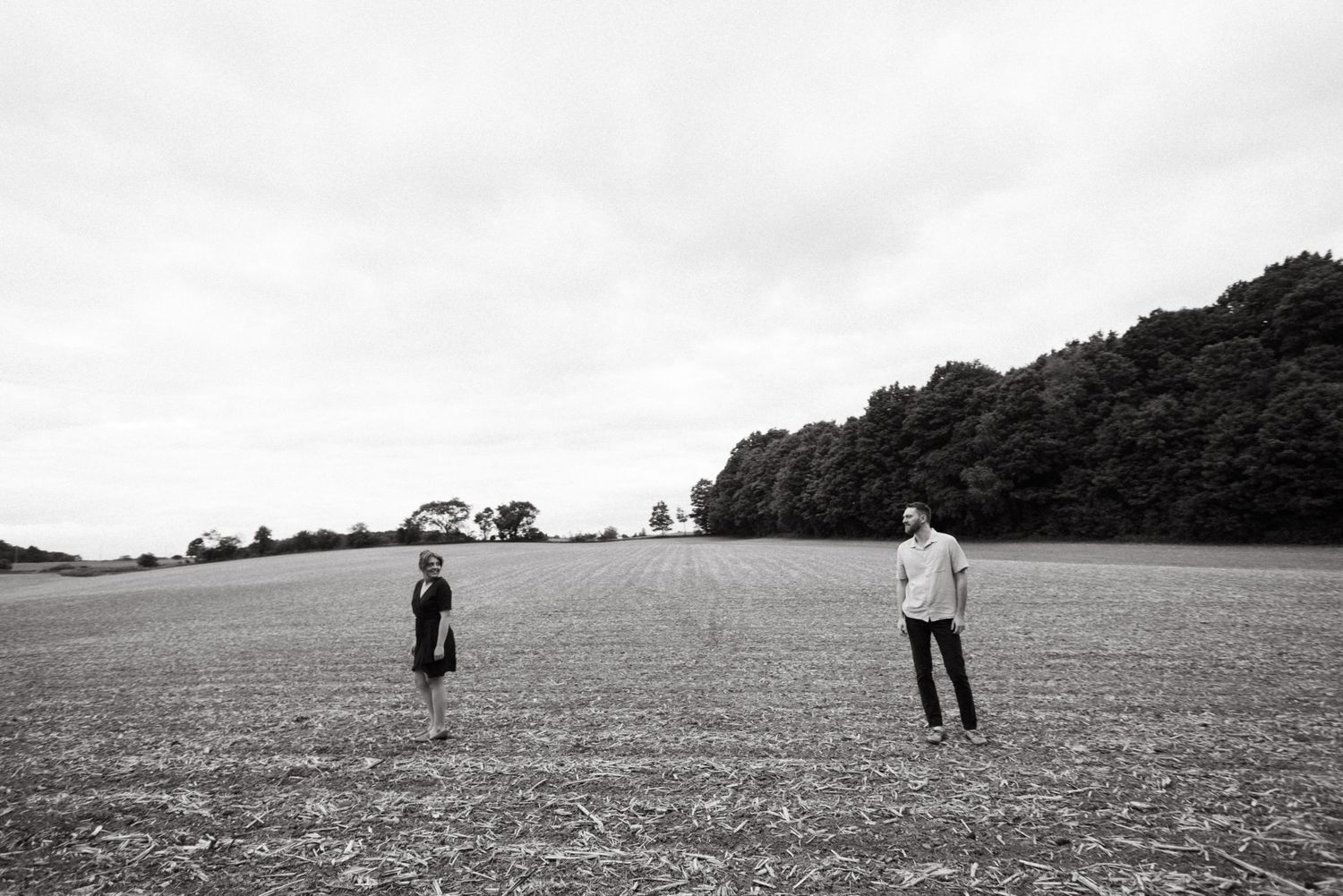 Two people stand at a distance from each other in a vast empty field with trees along the horizon in black and white taken in Guelph Ontario
