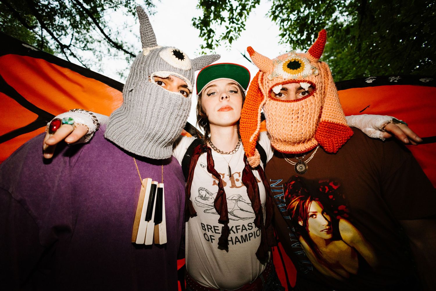 Three people pose together at a party wearing knitted monster masks and colorful clothing in a fun outdoor setting.