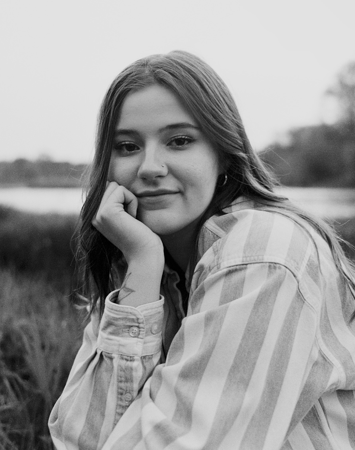 Black and white portrait photo of someone in a striped shirt sitting outdoors with hand resting on chin.