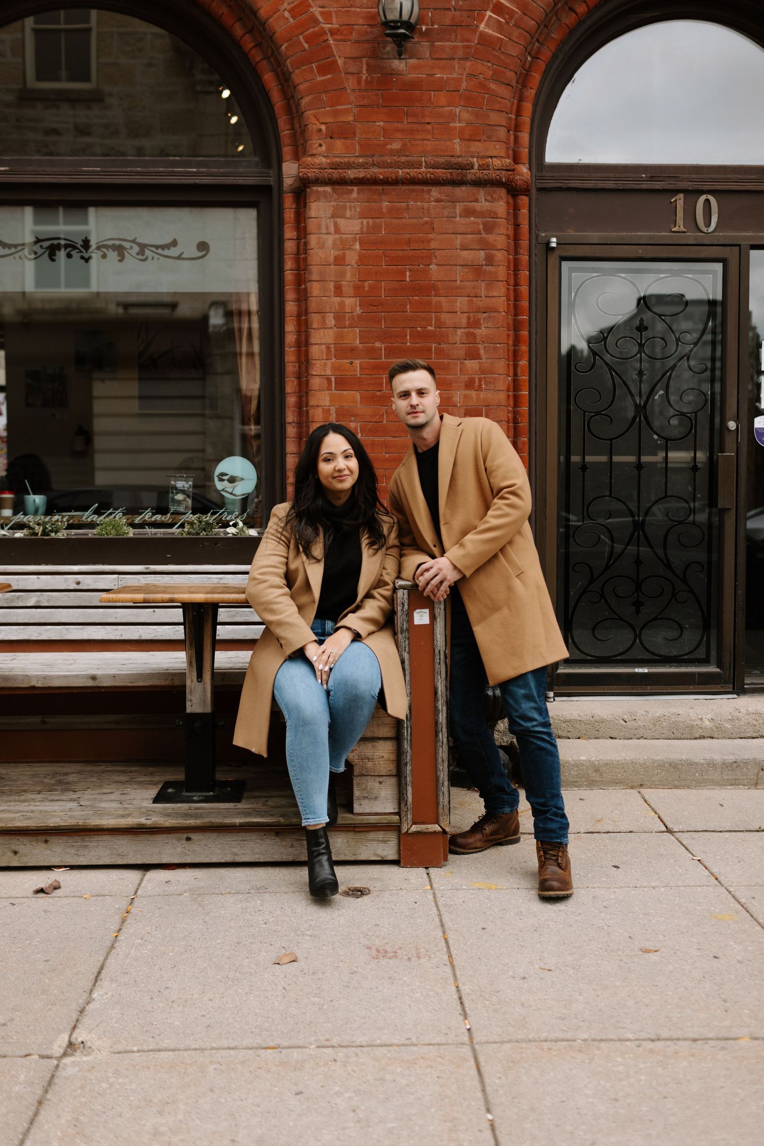 Two people wearing matching camel coats pose on a bench outside a brick storefront with a decorative metal door taken in Guelph Ontario