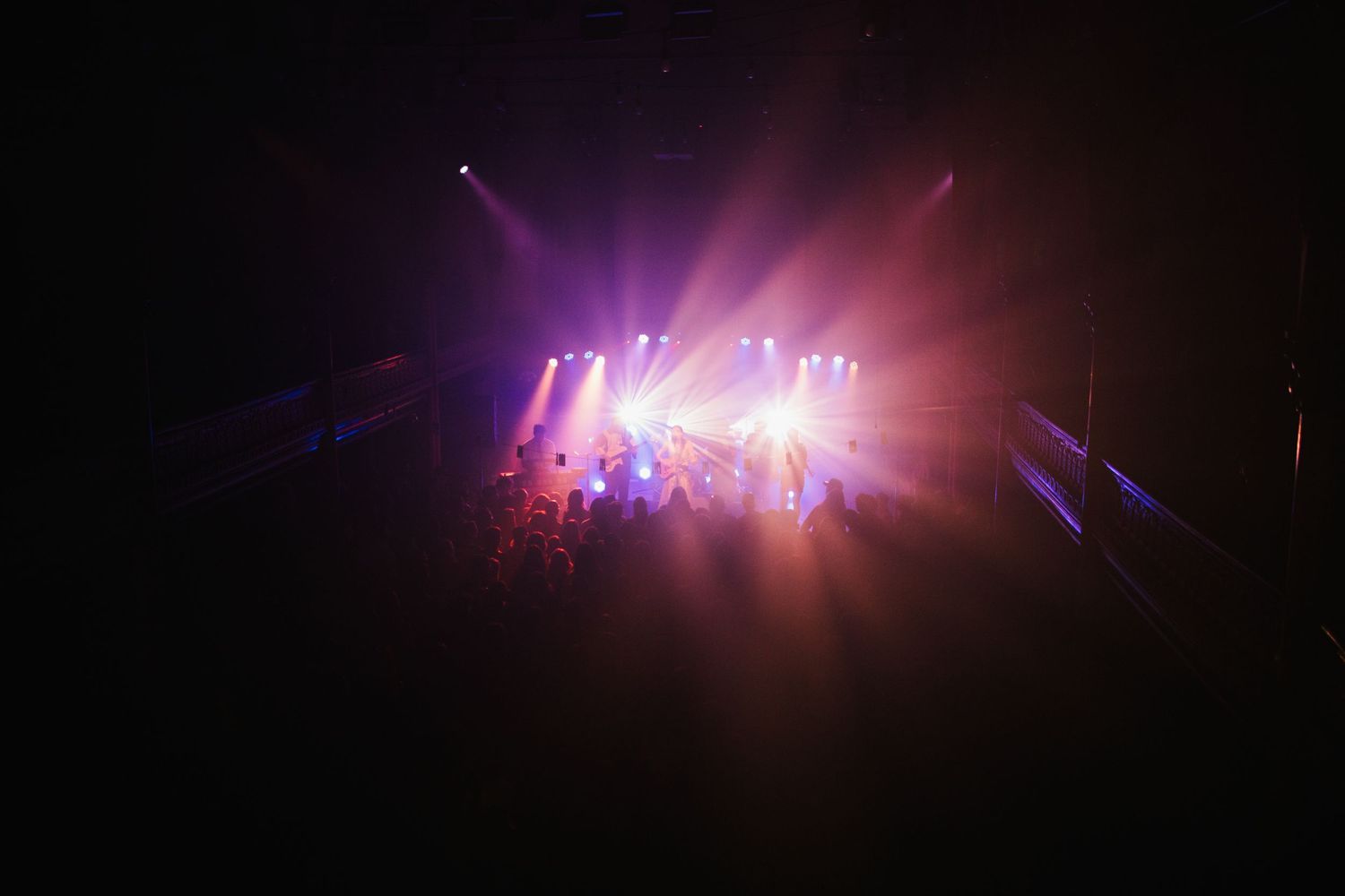 Colorful purple and pink stage lights illuminate a dark concert venue with bright beams cutting through the haze.