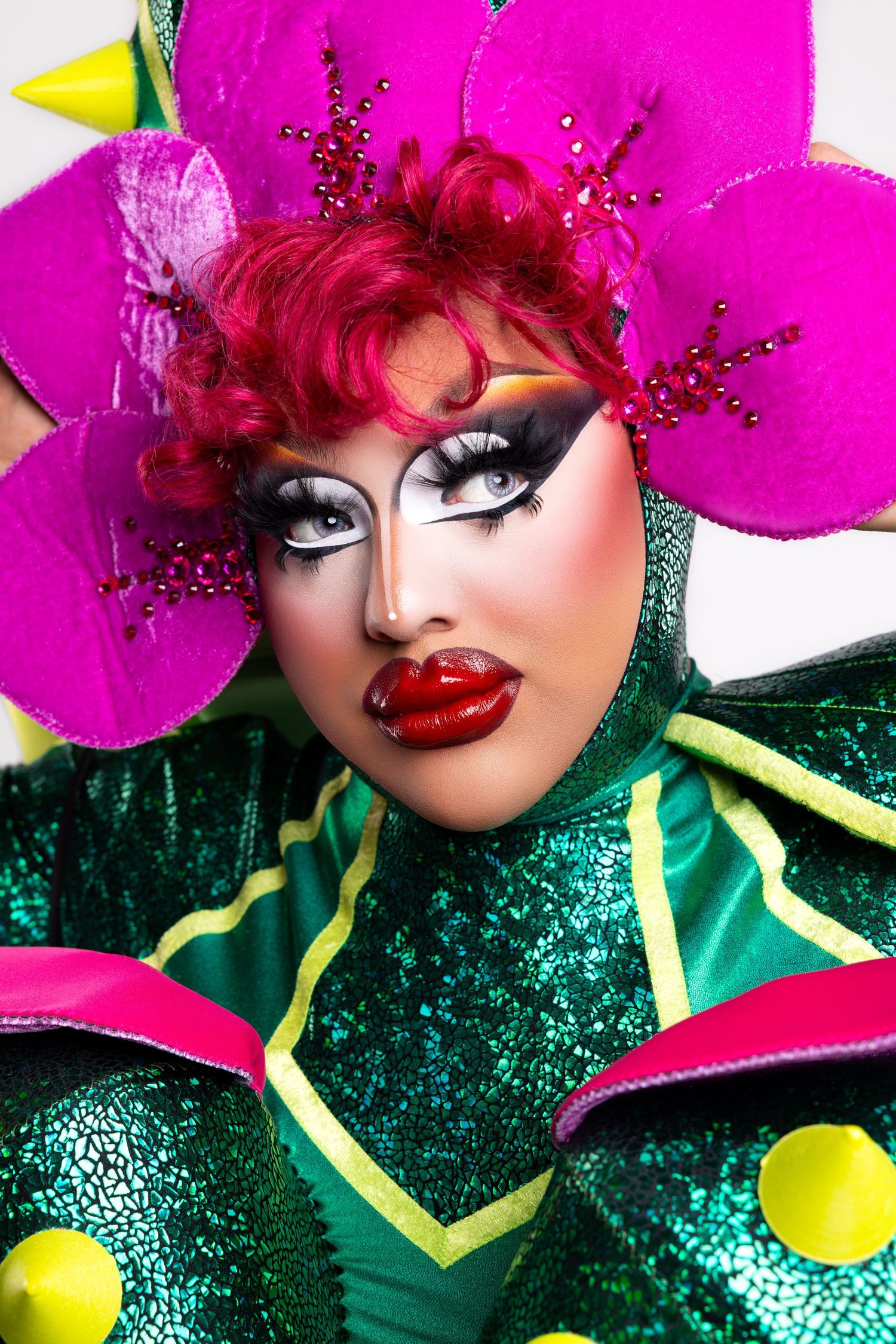 Vibrant drag performance look featuring bright pink flower petals, dramatic makeup, and sparkly green costume with yellow accents.