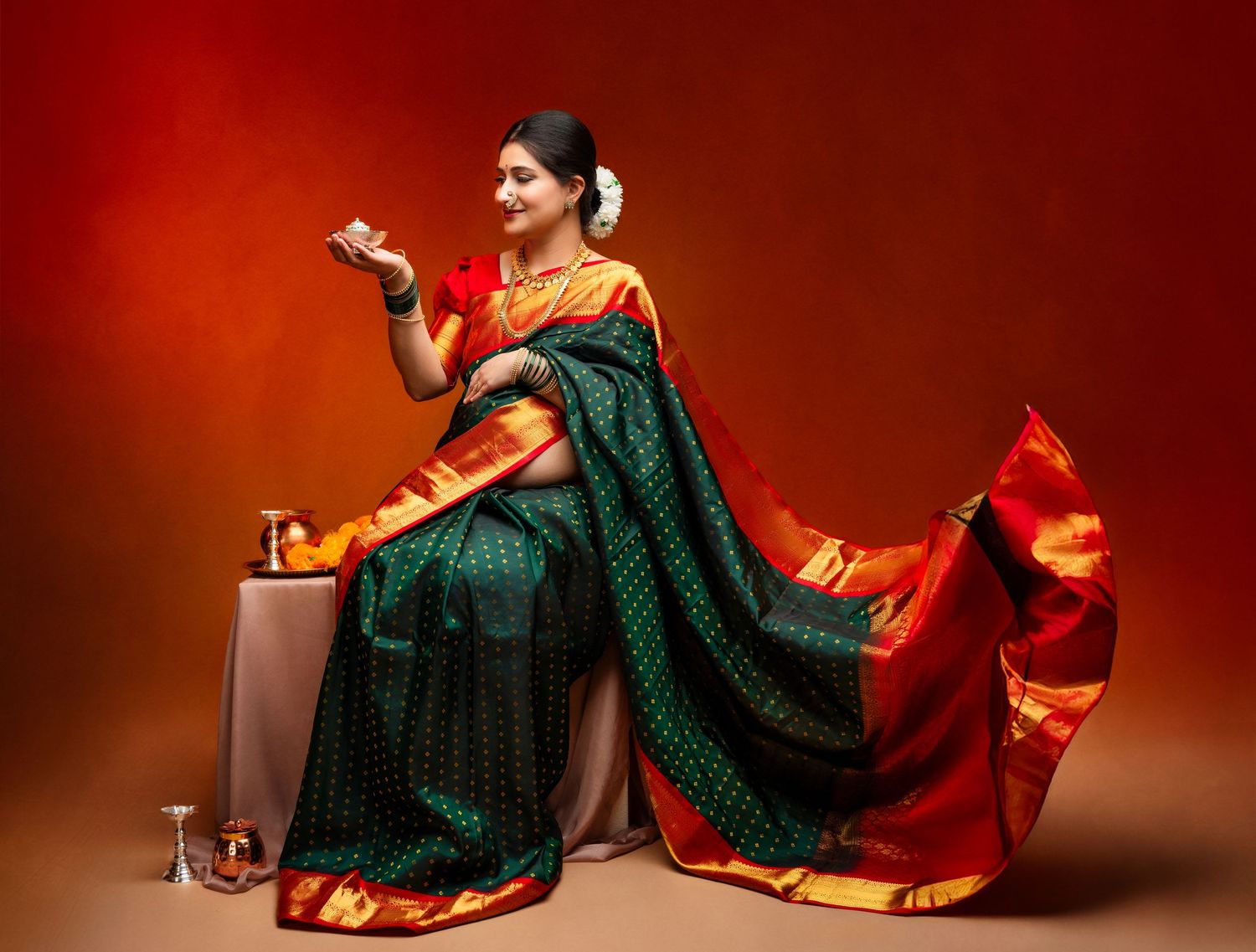 A traditional Indian saree maternity photoshoot with emerald green silk fabric and red accents against a dramatic red backdrop.