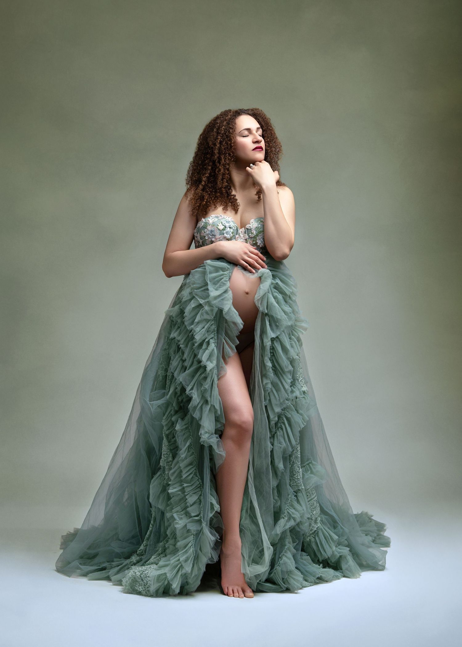 A glamorous studio portrait featuring a flowing sage green tulle gown with a dramatic high slit against a muted backdrop.