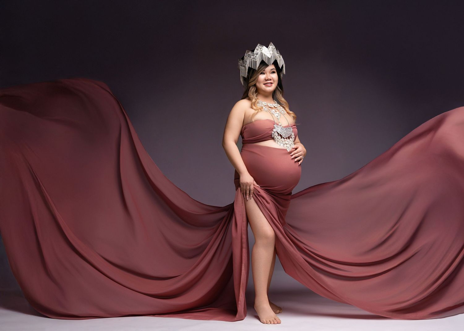 A maternity photo featuring flowing mauve fabric in a dramatic studio setting against a dark background.