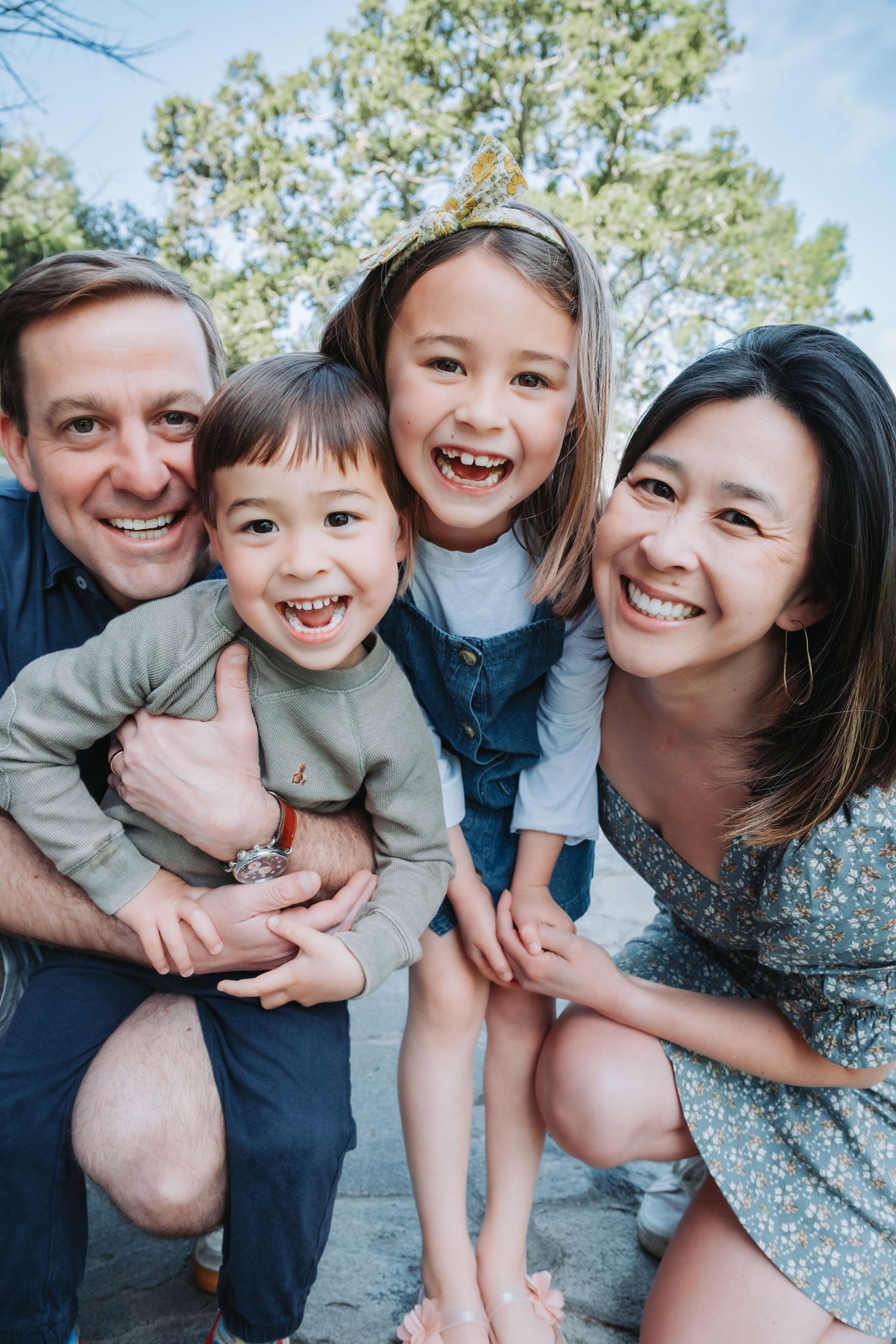 A happy interracial family with two young children sitting together outdoors with pine trees in the background.