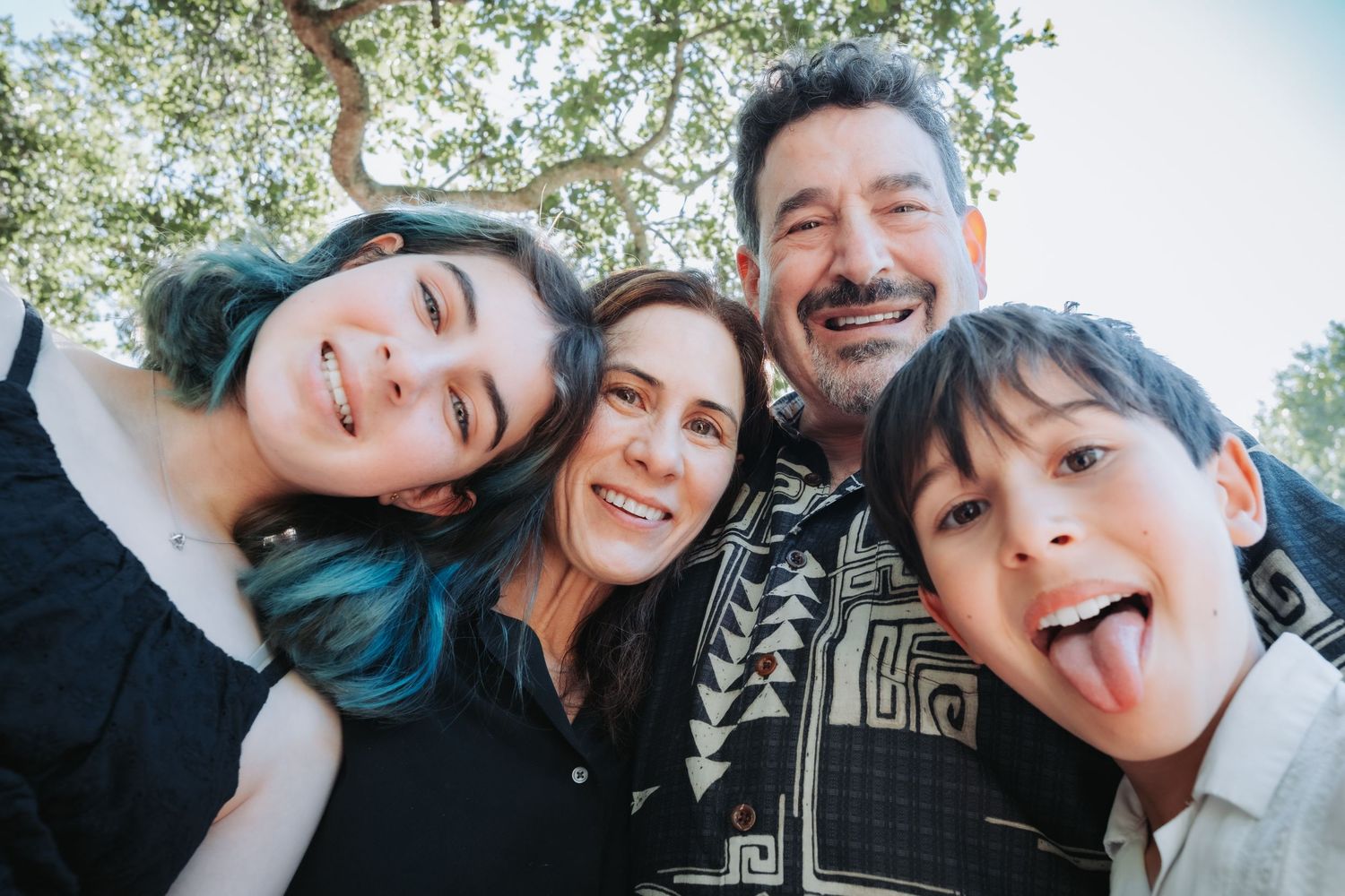 Group taking playful selfie photos outdoors against a backdrop of green trees on a sunny day.