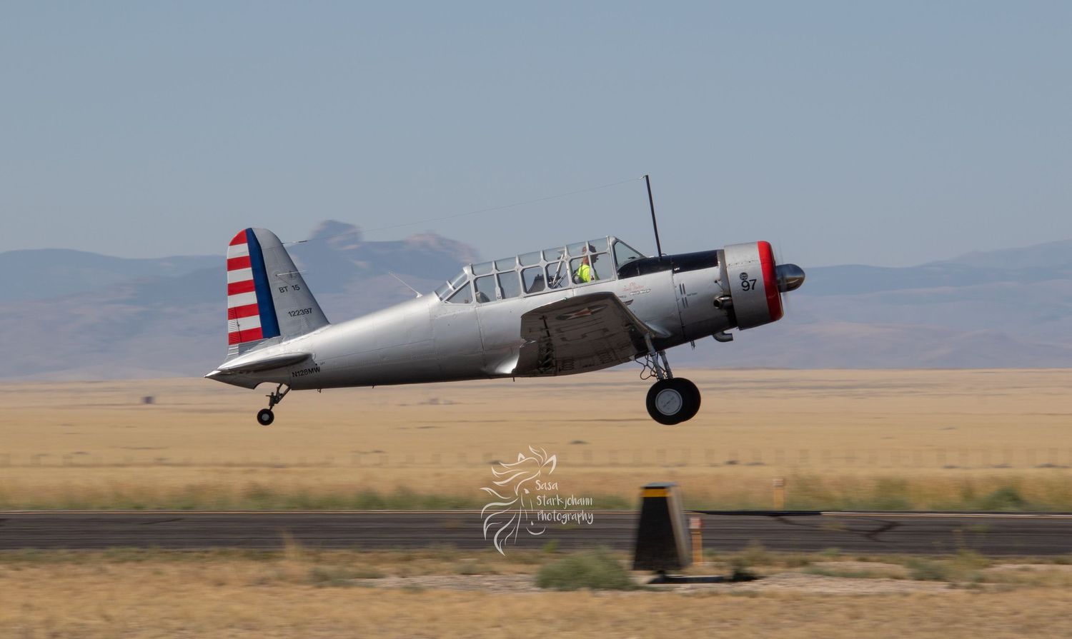 Vintage silver warbird aircraft with red and blue stripes taking off over desert landscape.