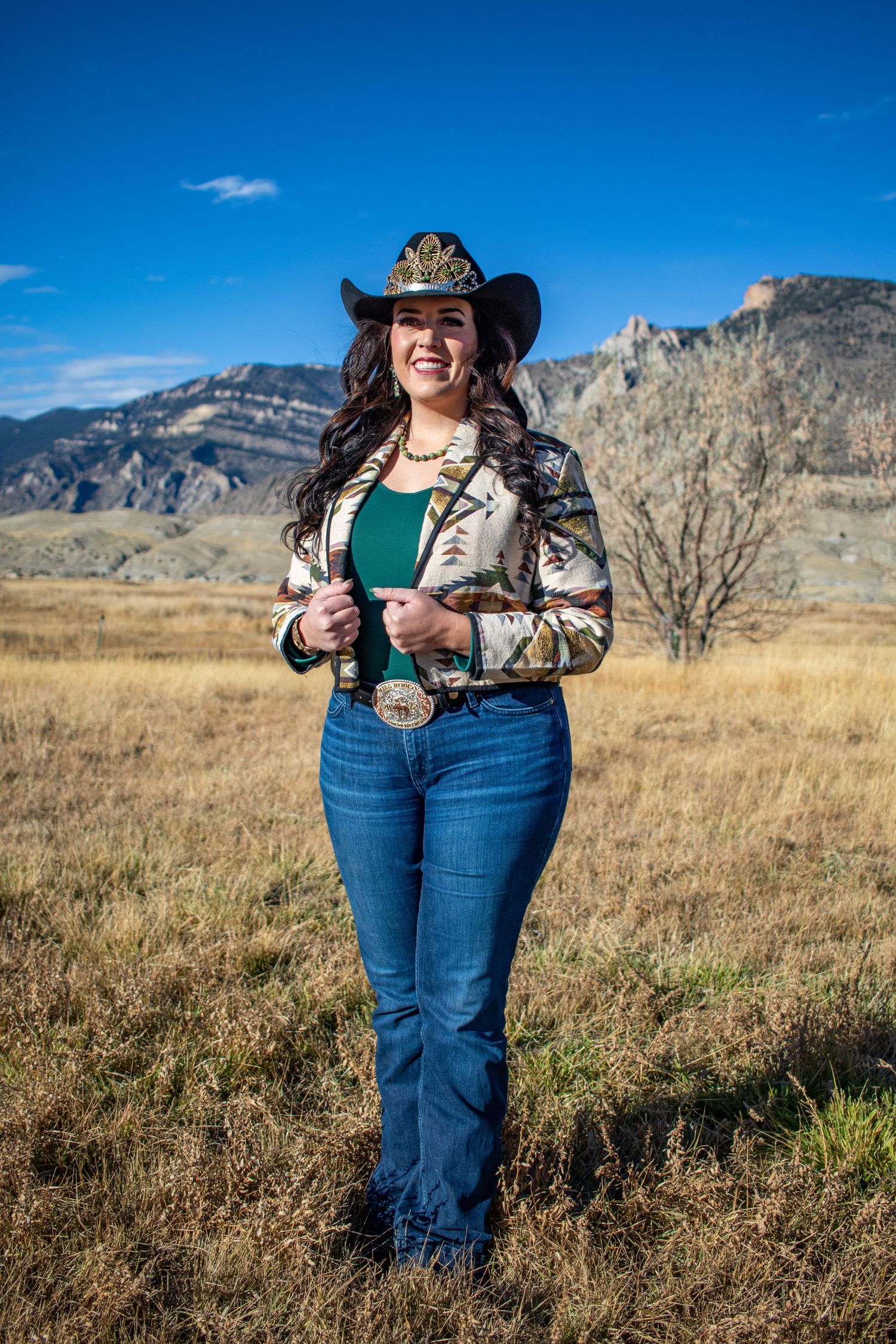 Western style portrait in outdoor setting with cowboy hat, belt buckle and casual attire against mountain backdrop.