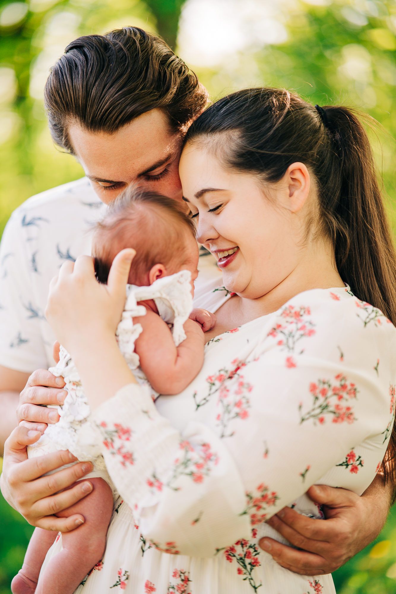 A loving couple shares a tender moment with their newborn baby outdoors in sunlit greenery.
