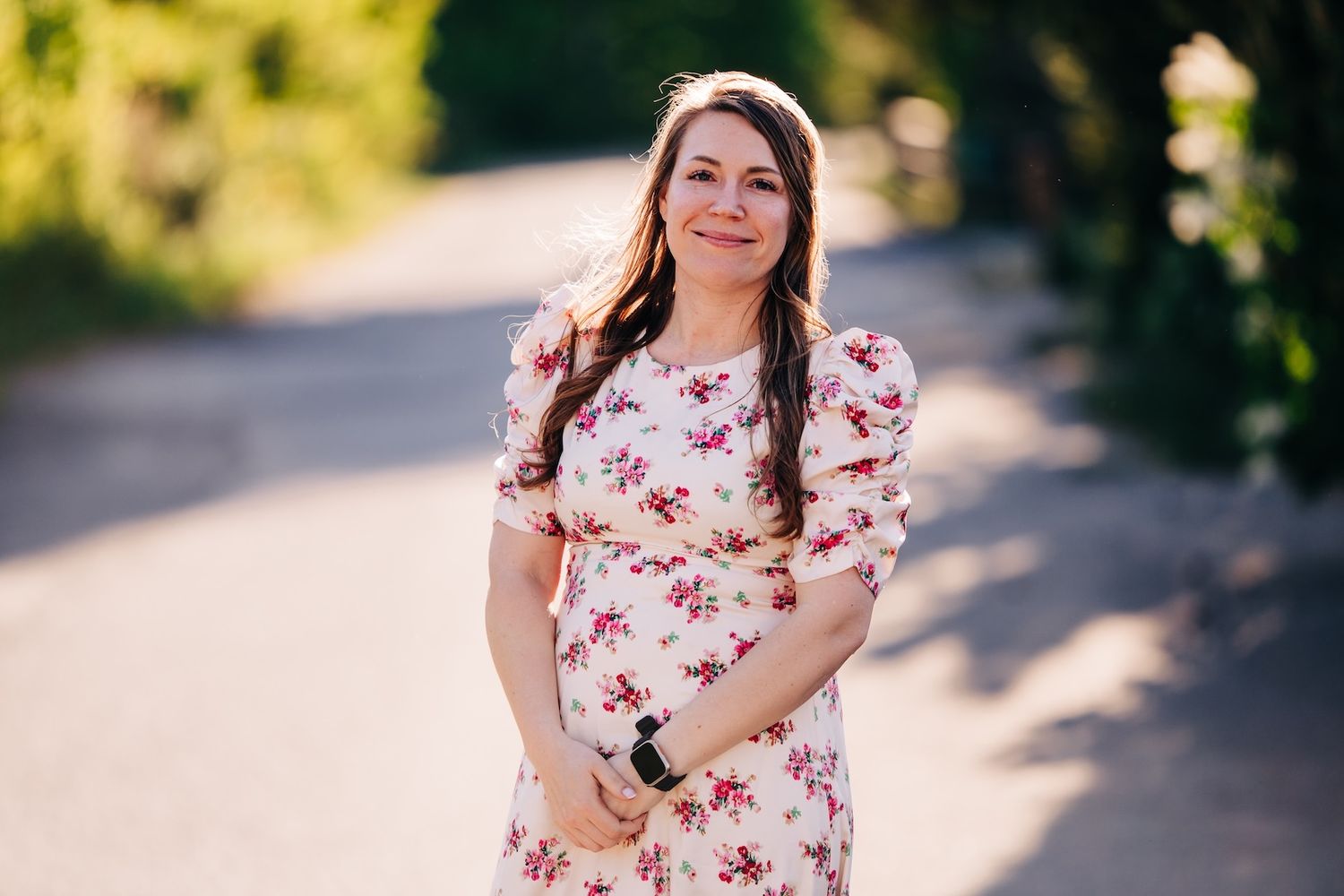 Expectant mother wearing floral pink dress stands on sunlit path outdoors during maternity photoshoot.