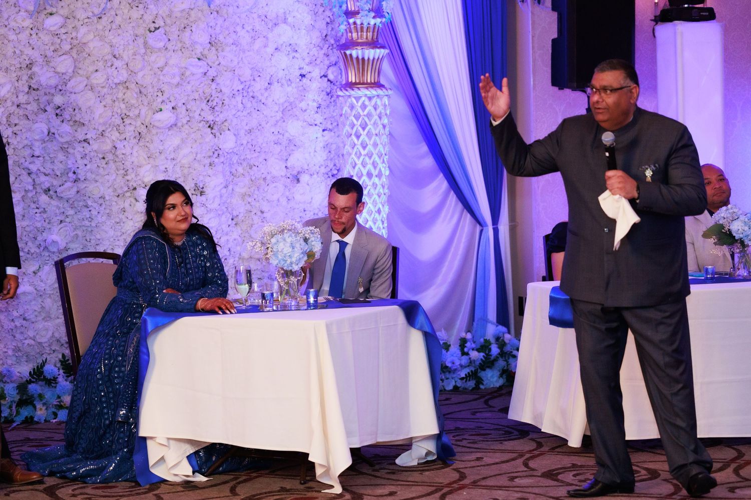 Wedding reception scene with guests at decorated table and speaker gesturing.