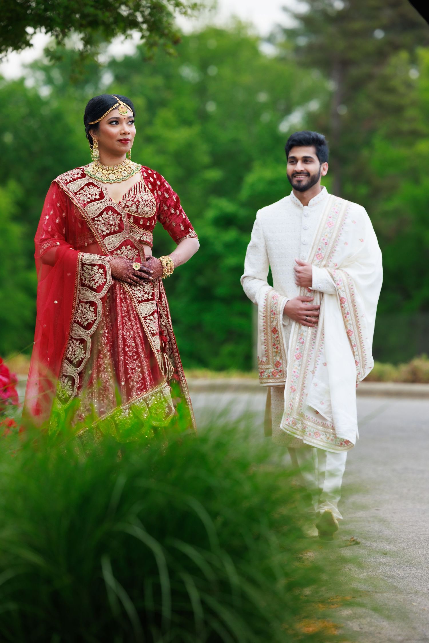 Couple in traditional Indian wedding attire posing on garden path with lush greenery.