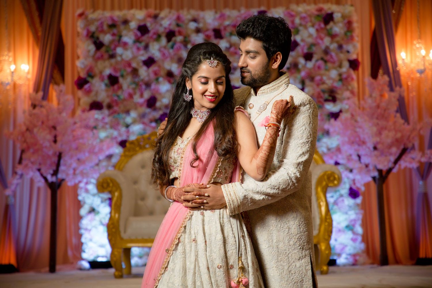 Couple in elegant Indian attire sharing a romantic moment against floral backdrop.