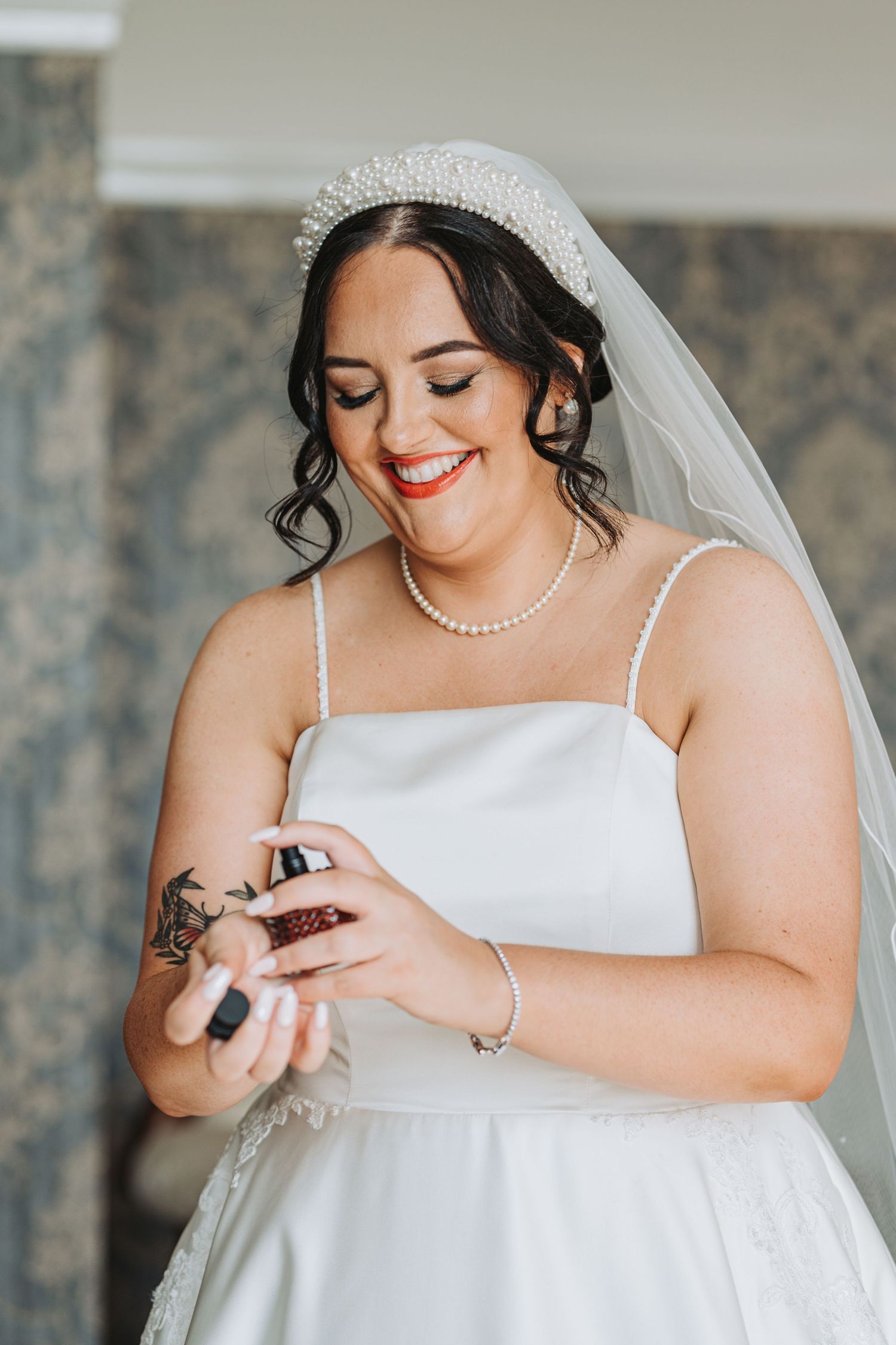 A bride in a white wedding dress with pearl necklace smiles while looking down at her hand on her wedding day at Oswald House.