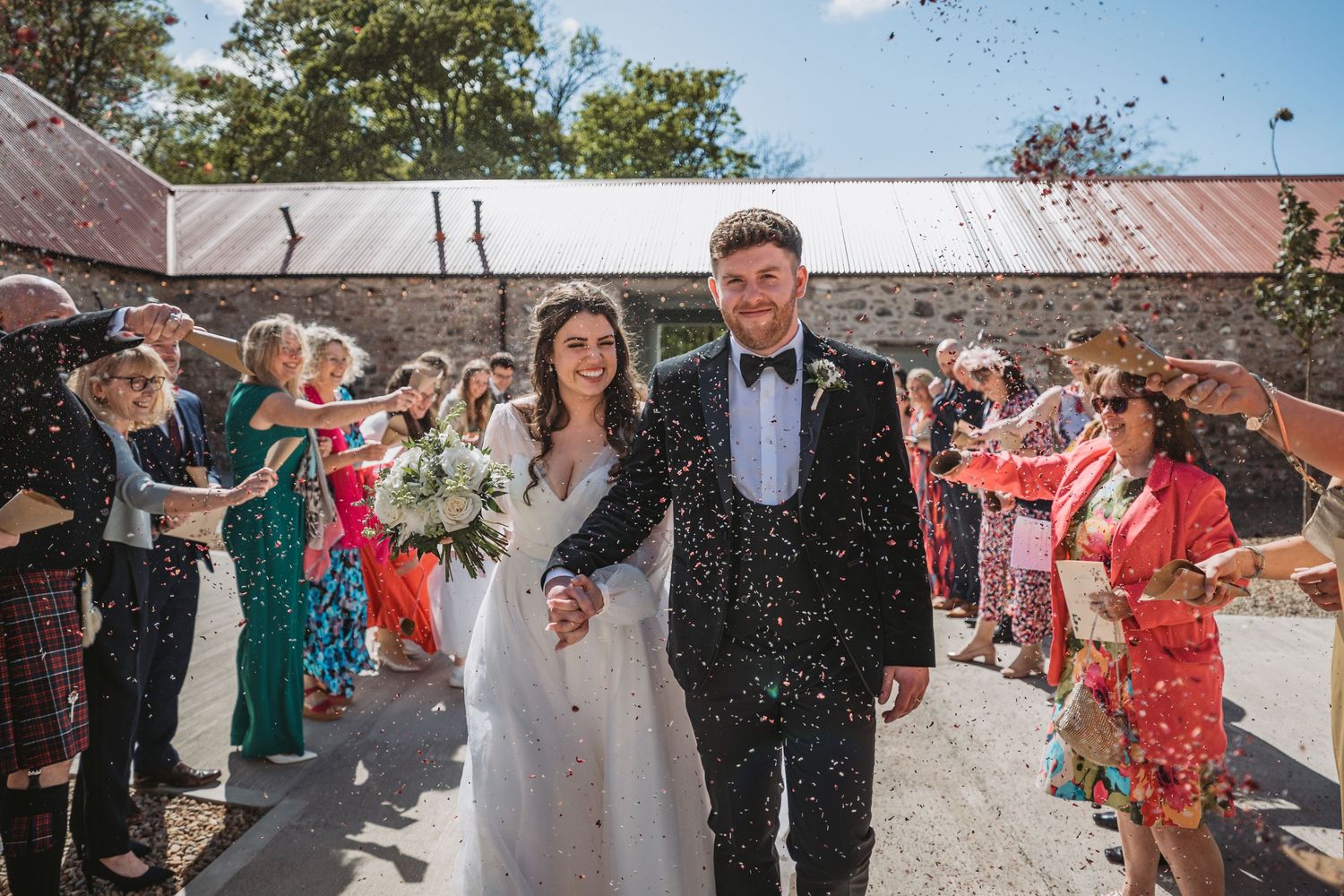 Wedding guests shower newlyweds with confetti as they exit Kinclune Steading during a joyful outdoor celebration.