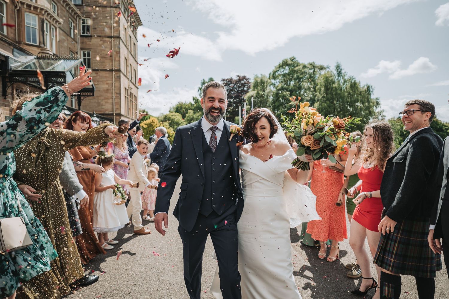 Newlywed couple walks through crowd of guests tossing flower petals at a wedding celebration outside Dunblane Hydro.