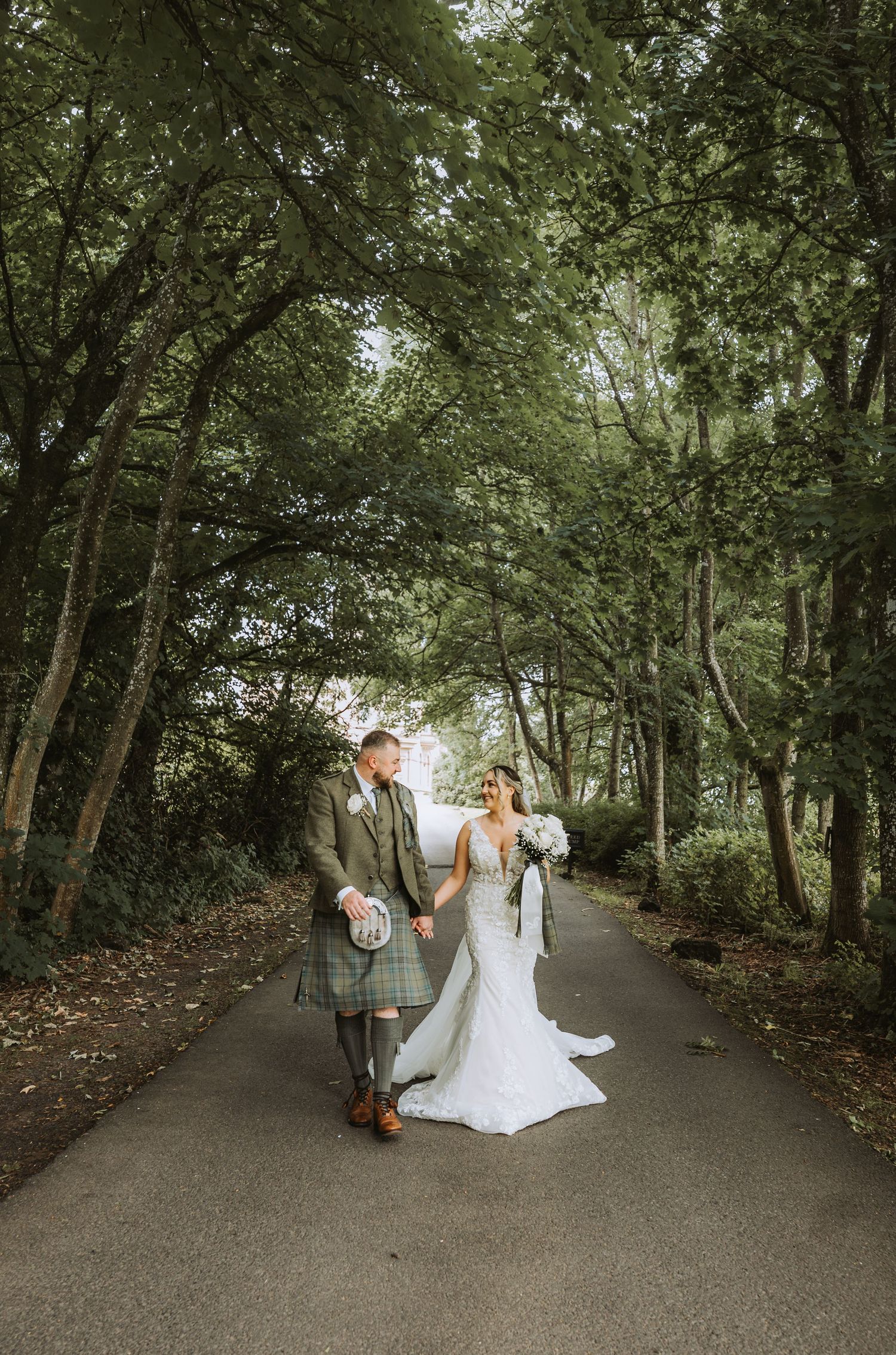 A wedding couple walks together down a tree-lined path at Cornhill Castle on their special day.