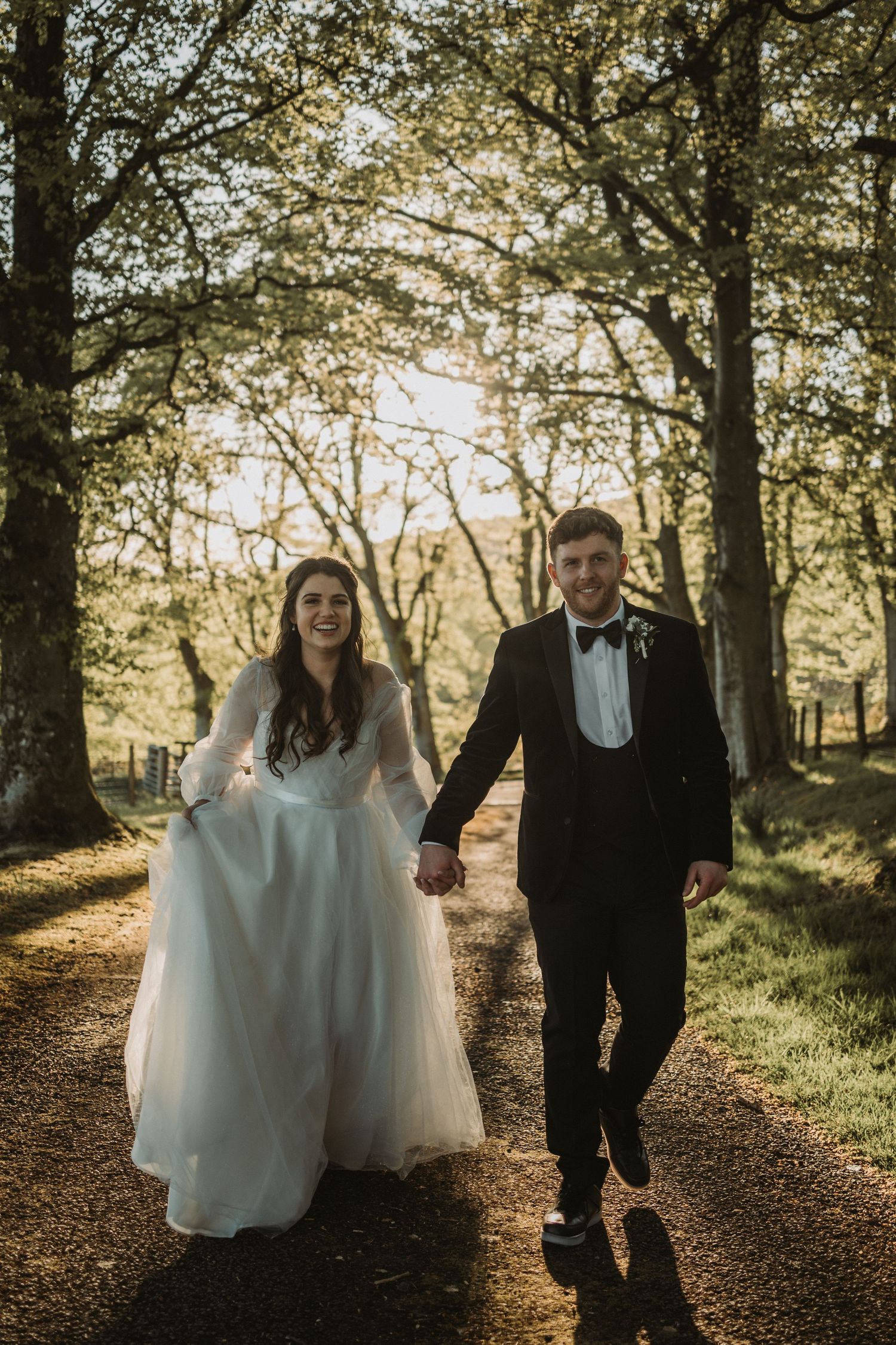 A couple walks hand in hand through a sunlit forest path at Kinclune House in wedding attire.