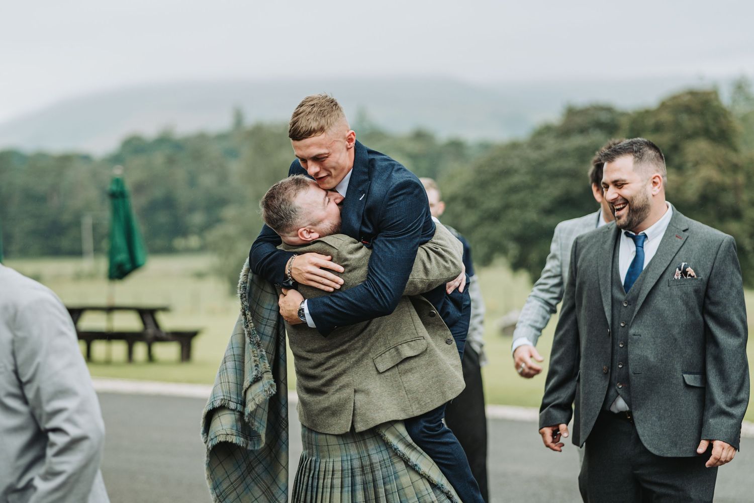 Two people share a joyful embrace while wearing traditional Scottish kilts as others look on at an outdoor gathering at Cornhill Castle.