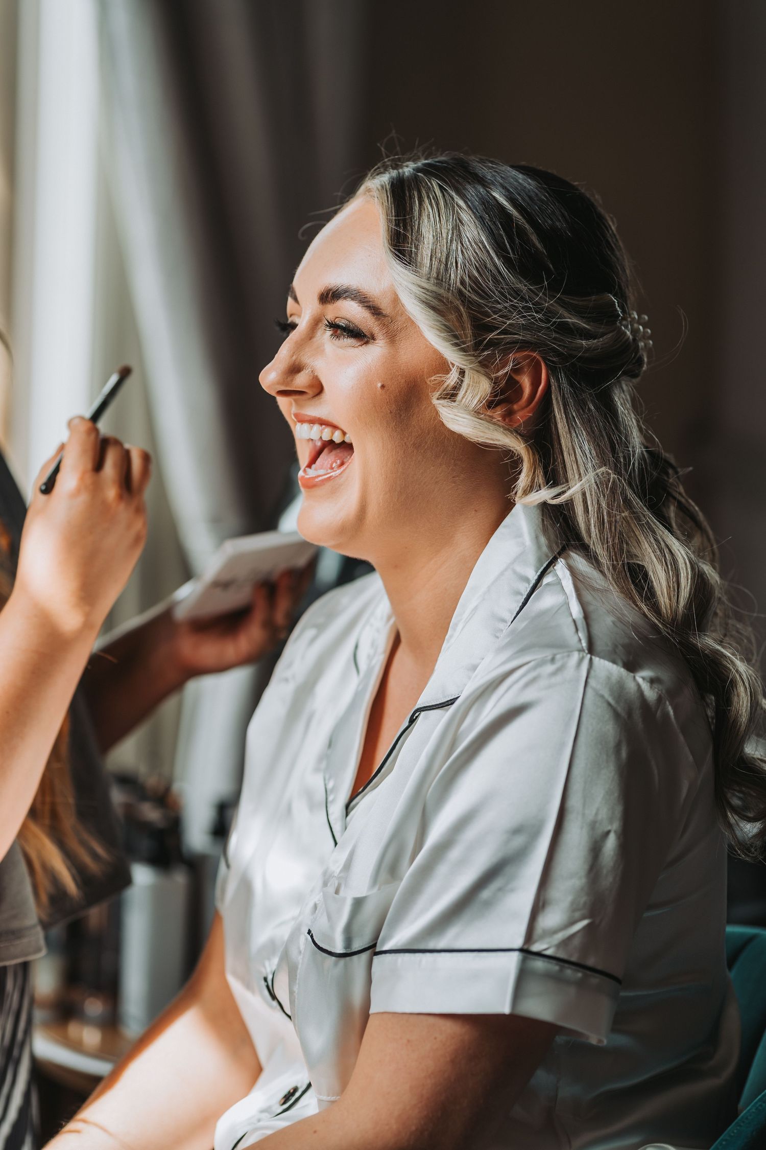 Someone laughs joyfully while getting their makeup done, wearing a white satin robe in natural window light at Cornhill Castle.