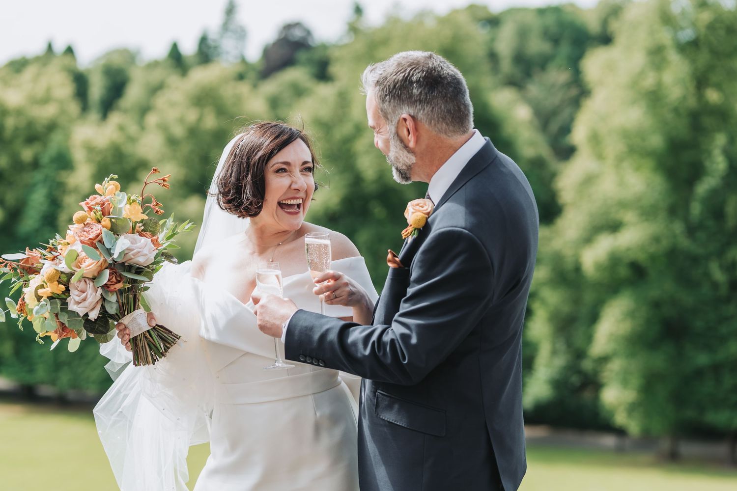 Joyful bride in white dress laughs while holding colorful bouquet outdoors with greenery of Dublane Hydro in background.