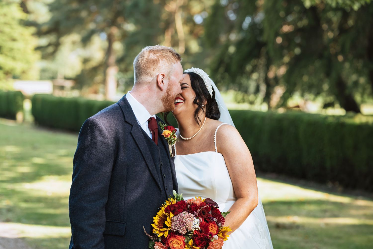 A joyful wedding couple share an intimate moment outdoors while holding a vibrant sunflower and burgundy bouquet.
