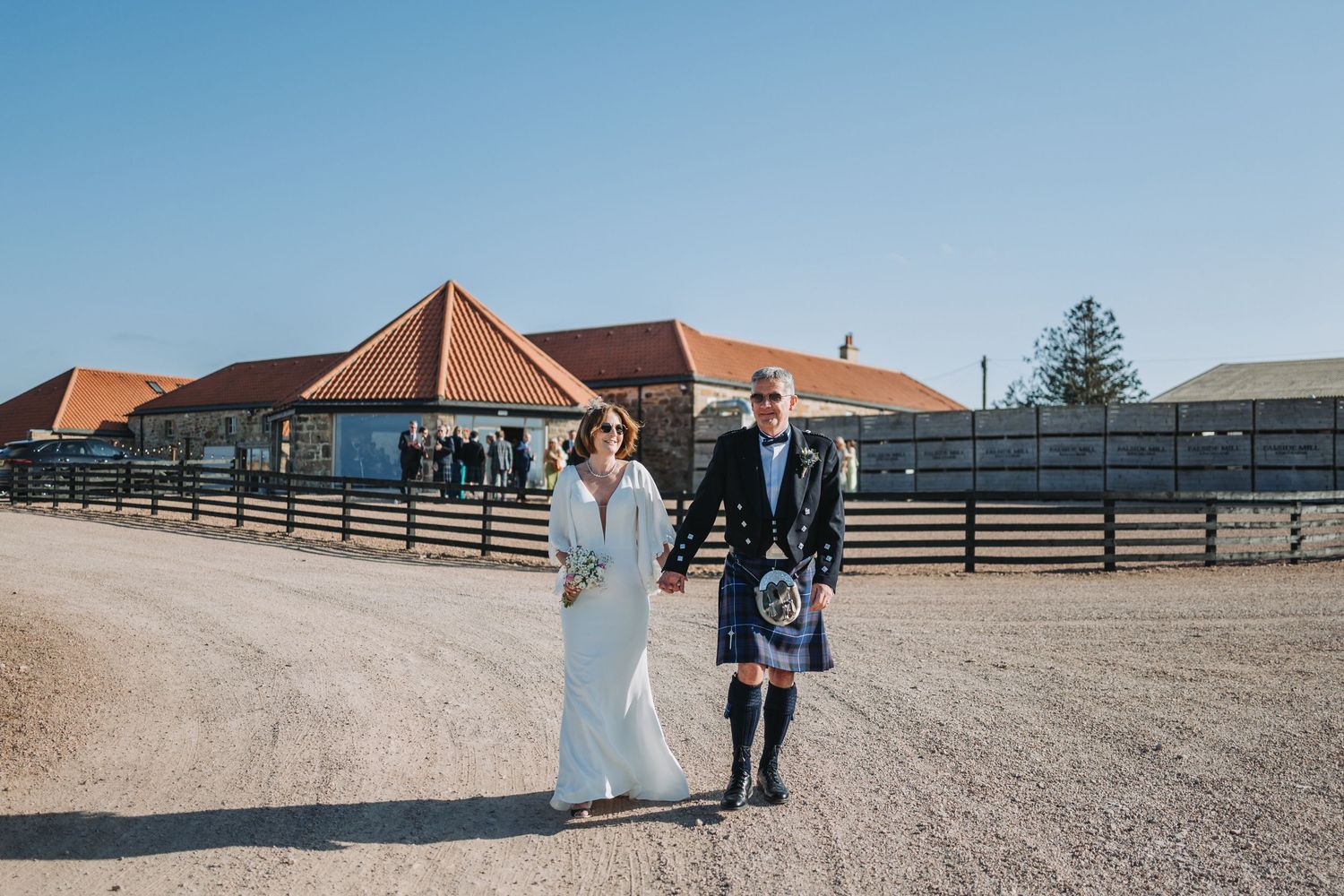 A couple walks together on a dirt path outside rustic barn venue Falside Mill with wooden fences in the background.