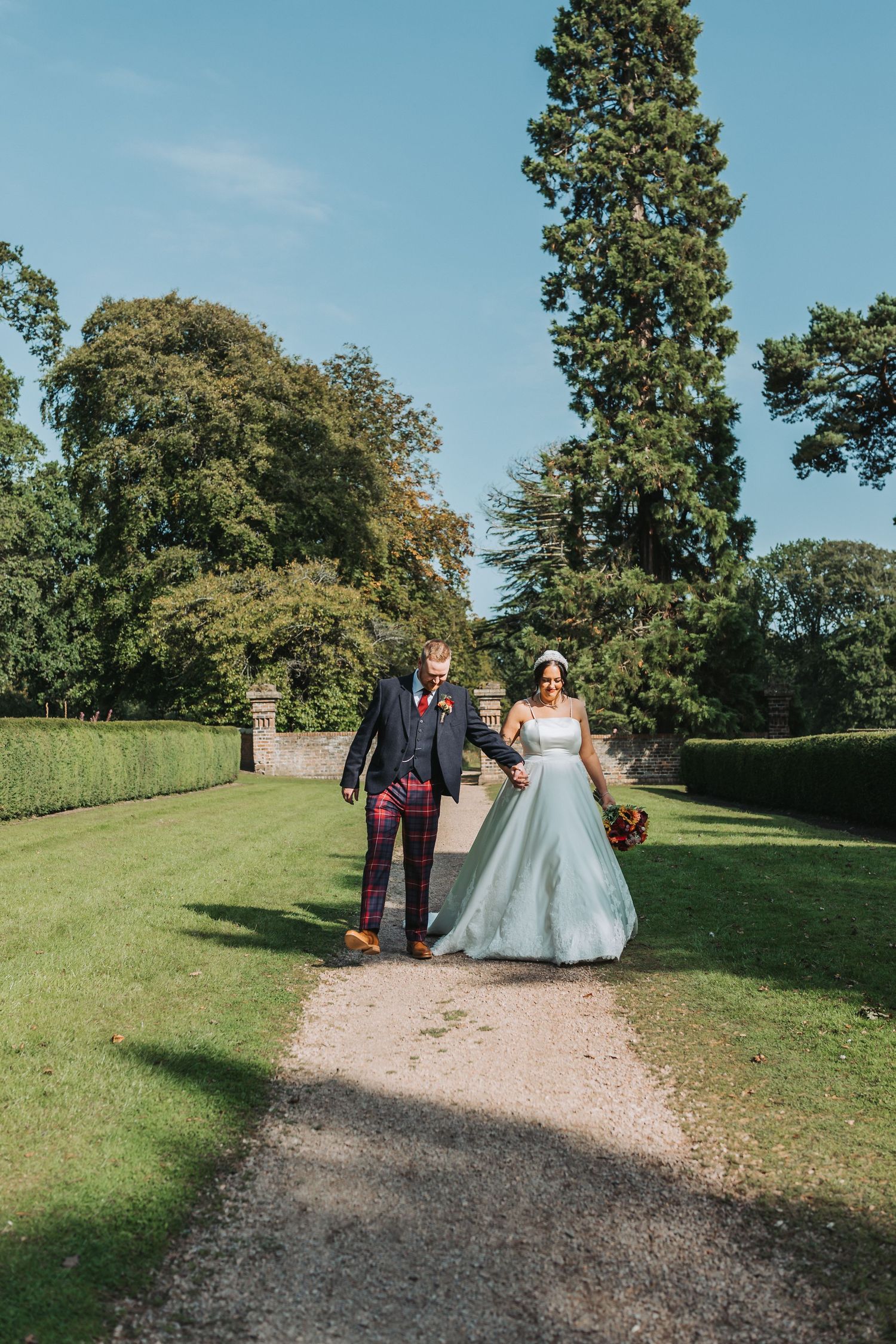 A couple walks hand in hand down a gravel path between manicured lawns and tall trees at elegant garden venue Oswald House.