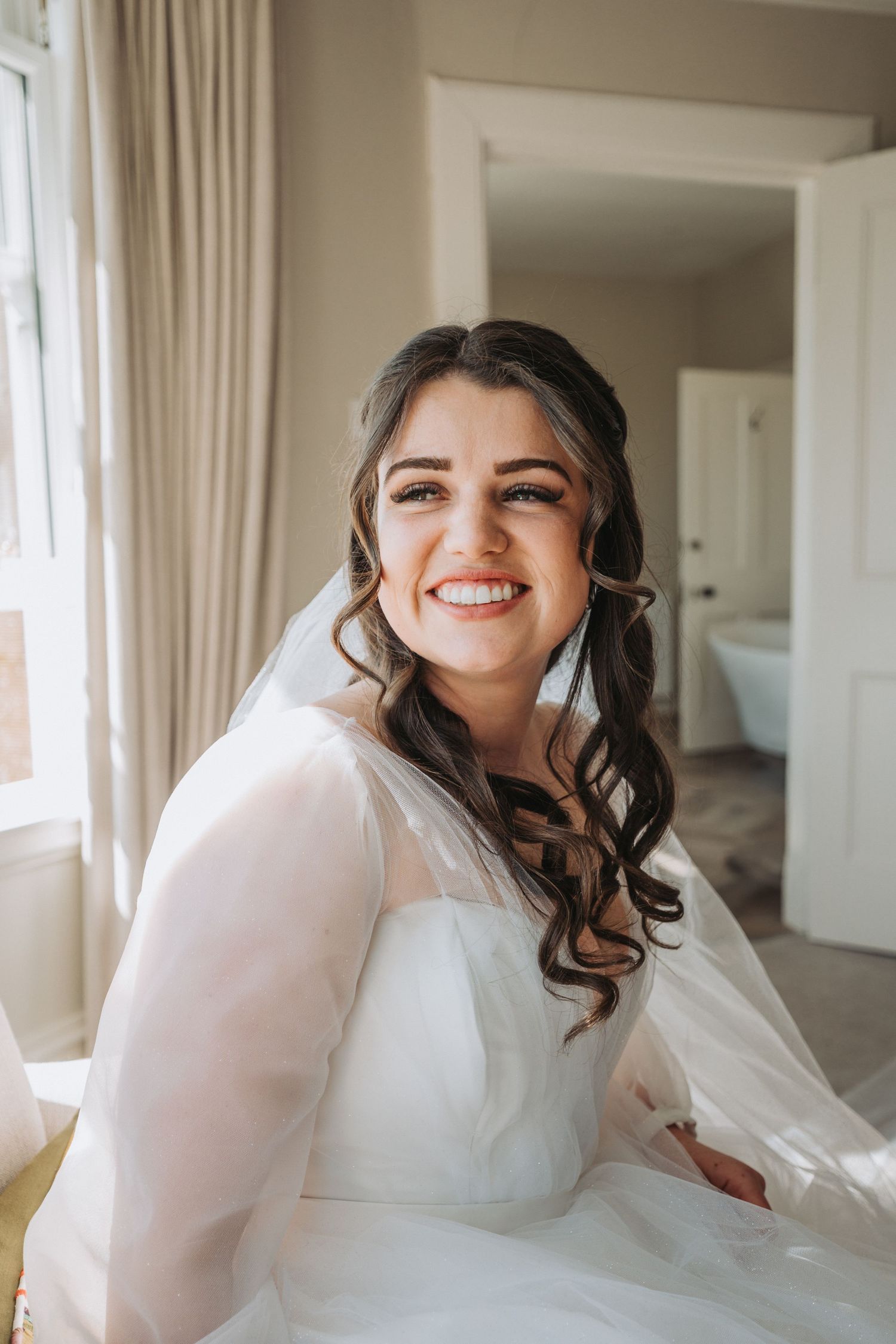 A bride in a long-sleeved white wedding gown with sheer details poses by a window with flowing curled hair.