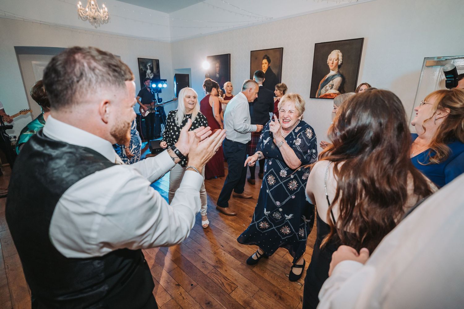 People enjoying a lively dance party on a wooden floor in a room with portrait paintings on the walls at Oswald House.