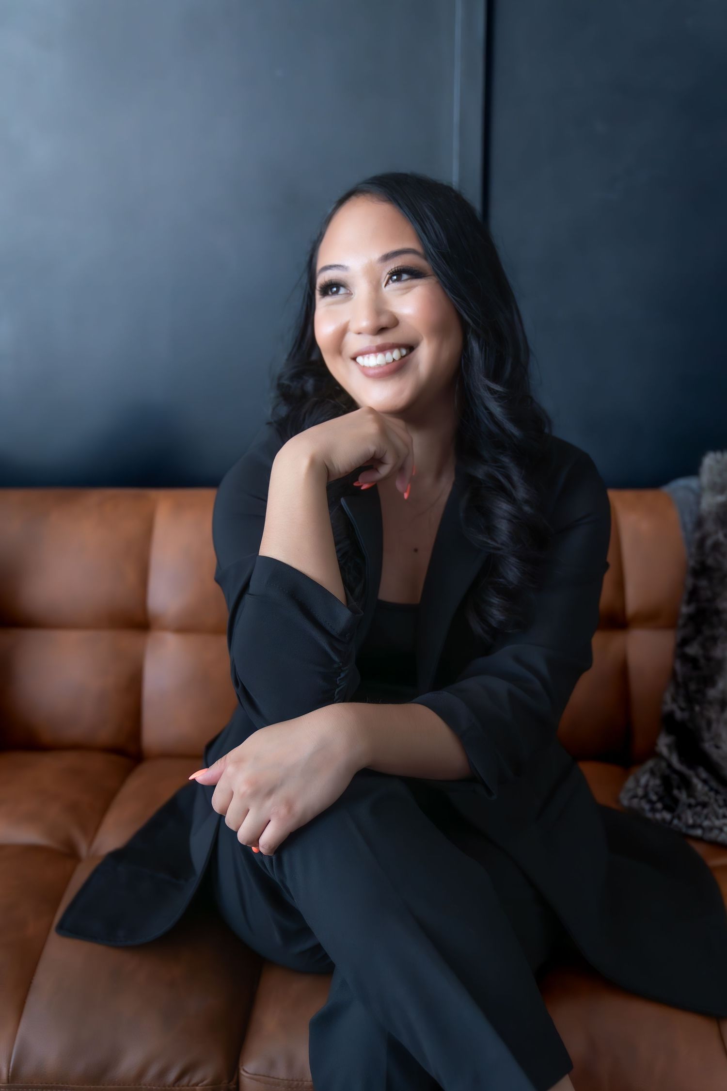 girl wearing a black suit, sitting on a brown couch smiling.