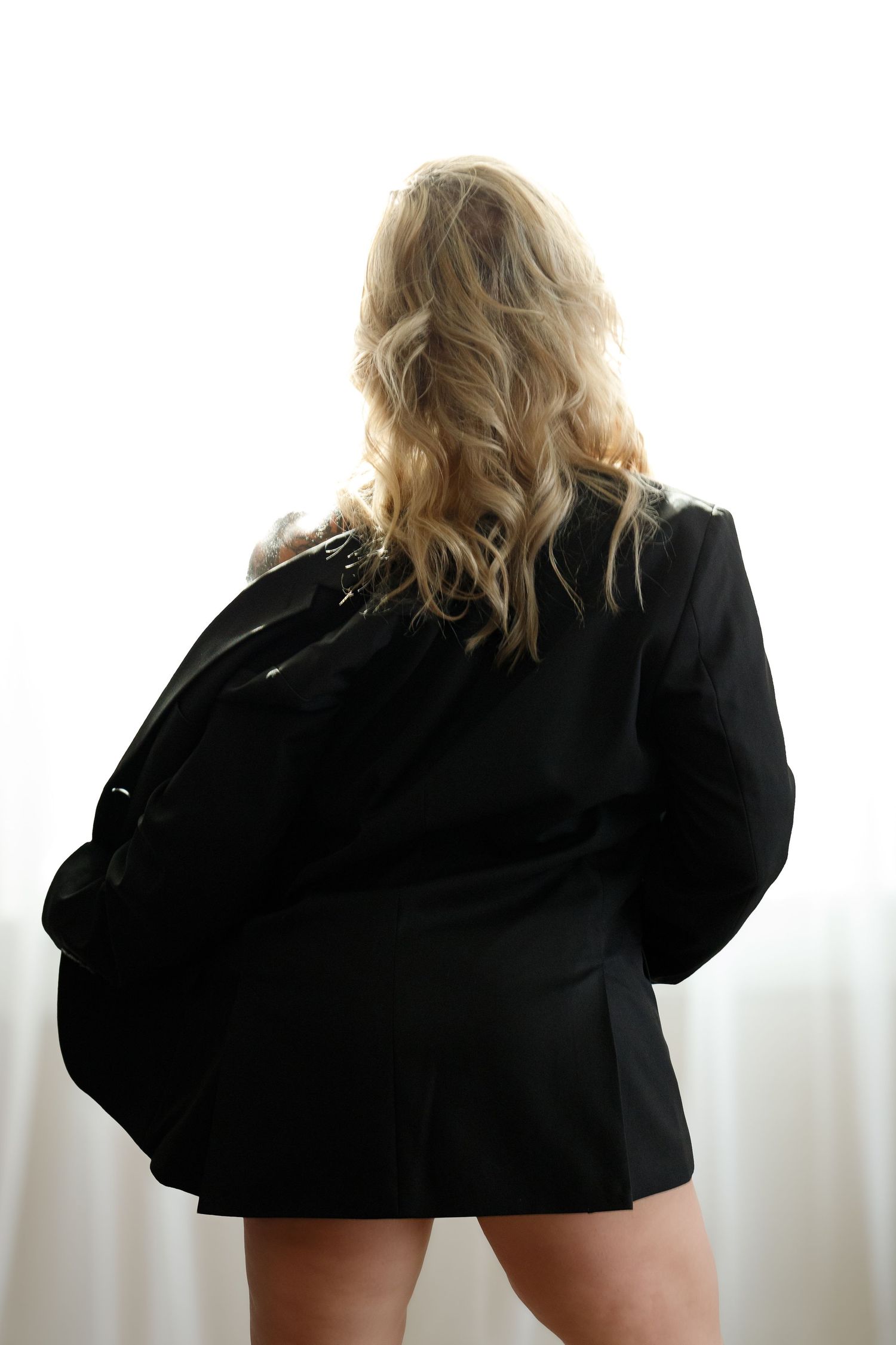 Back view of a person with wavy blonde hair wearing an oversized black blazer against a white background.