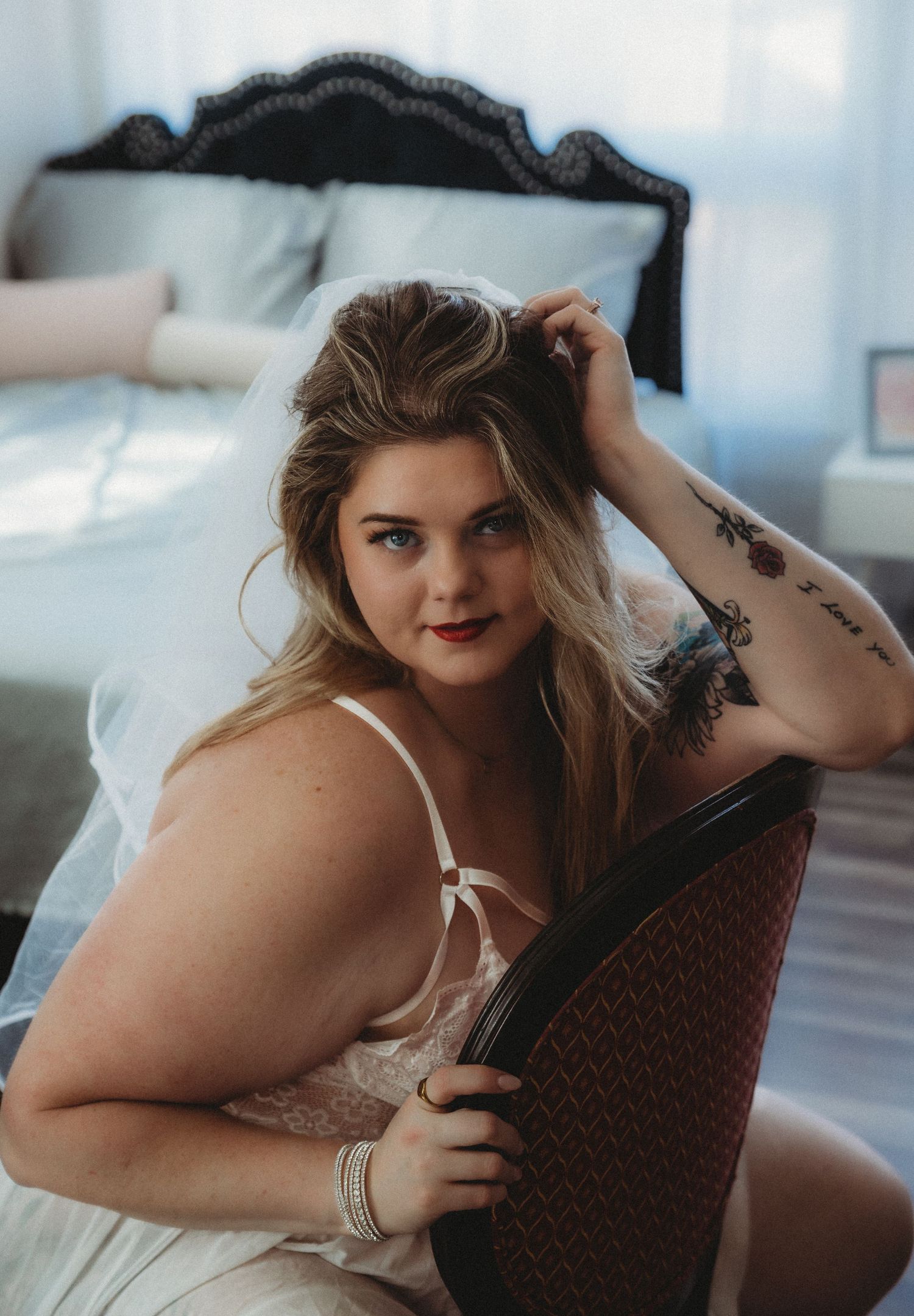 Lounging in a white top and bedding, a model with tattoos poses on furniture in soft natural lighting.